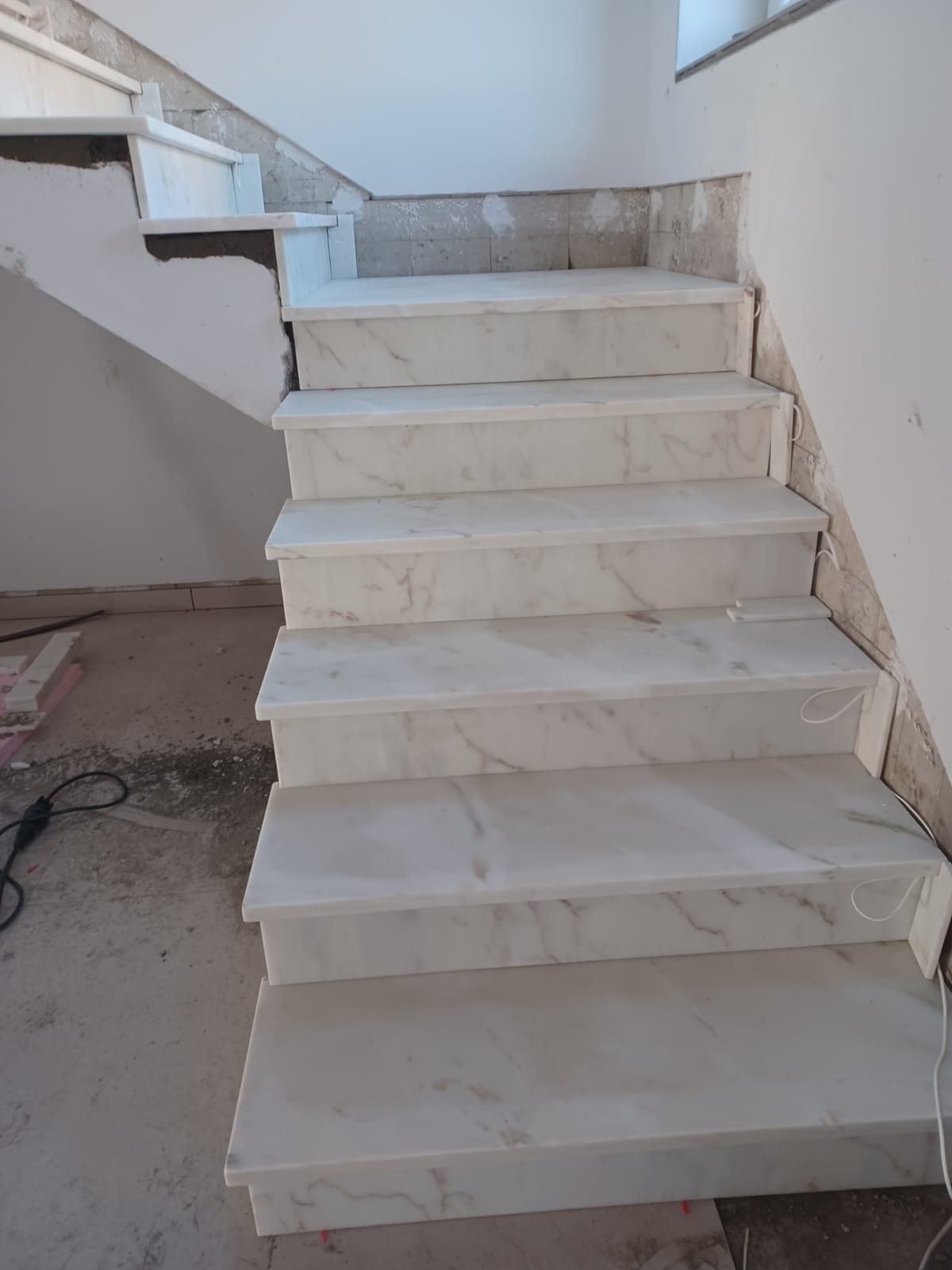 Wykończone marmurowe schody wewnętrzne w trakcie montażu, widoczne niedokończone ściany i podłoga. Jasne, naturalne światło podkreśla wzór kamienia.