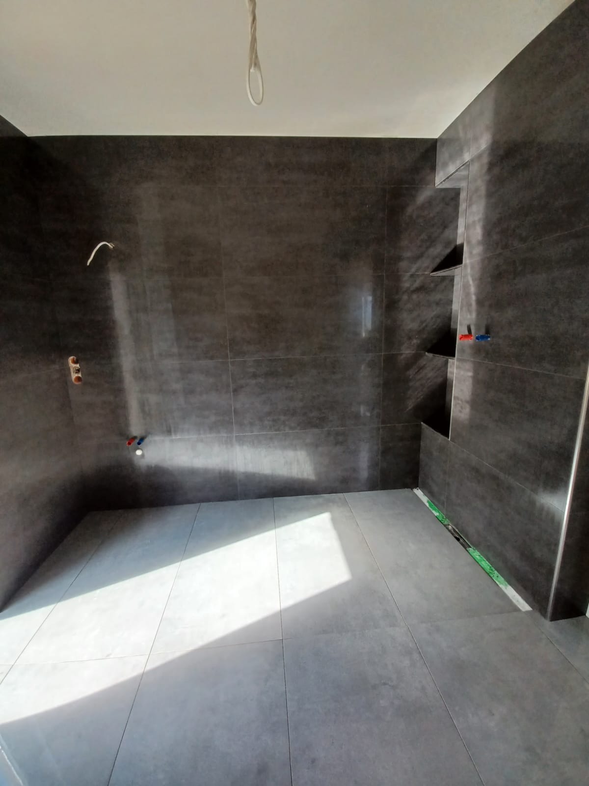 Wykończona łazienka z ciemnymi płytkami na ścianach i jasnymi na podłodze. Widoczne półki wnękowe i instalacja wodna. Minimalistyczny design, nowoczesne oświetlenie.