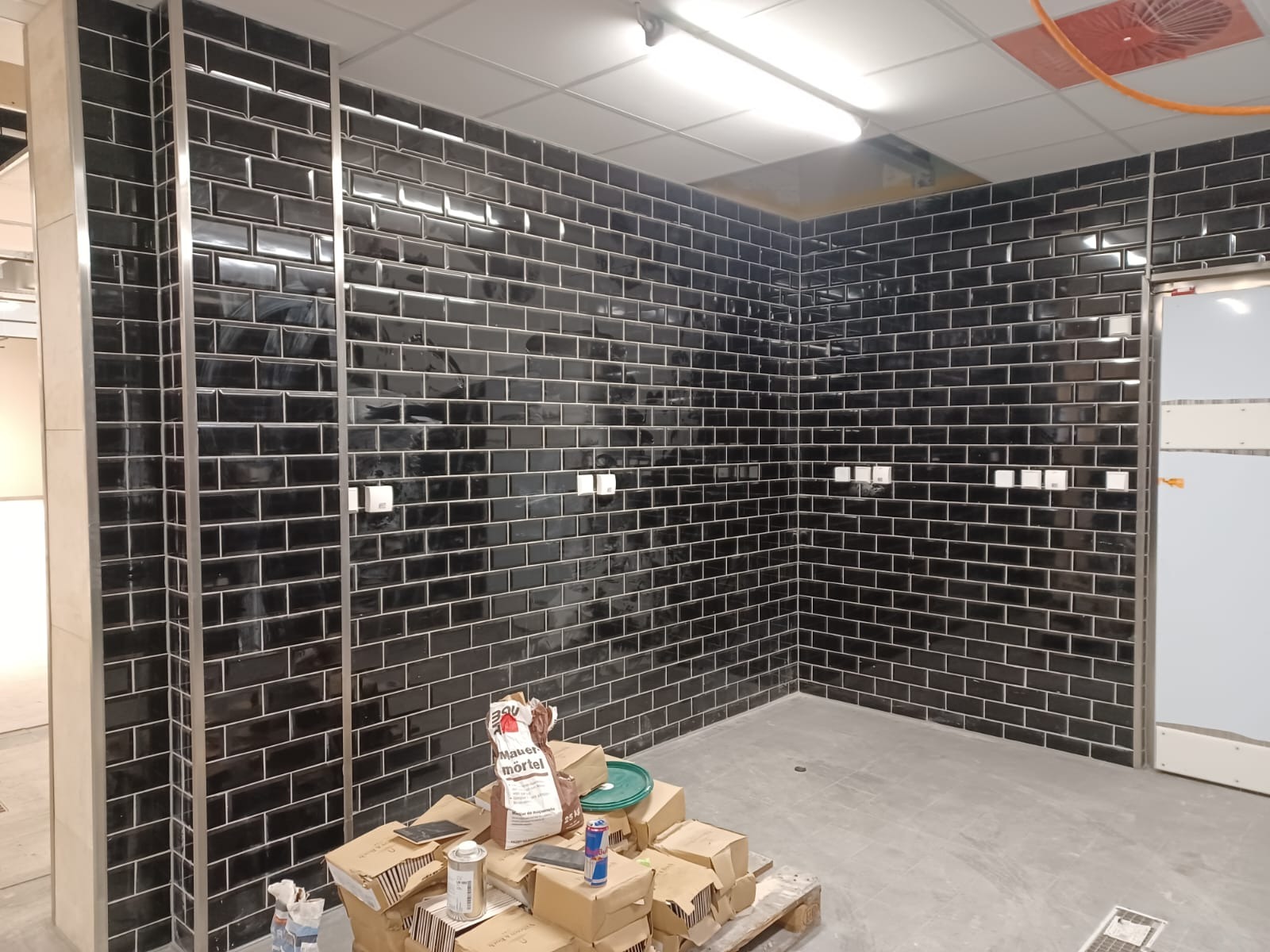 Czarne, błyszczące płytki ułożone w cegiełkę na ścianach. Widoczne puszki elektryczne, materiały budowlane na palecie i fragment sufitu z oświetleniem.