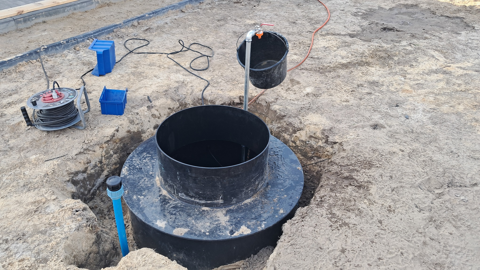 Czarna studnia z wodą w wykopie, widoczne narzędzia i akcesoria. Ujęcie z góry, ukazujące etap prac przy instalacji studni.