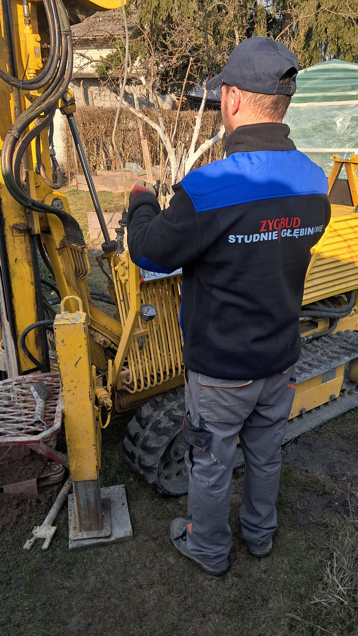 Pracownik w firmowej bluzie obsługuje żółtą wiertnicę do studni głębinowych. Widoczne detale maszyny i otoczenie placu budowy.