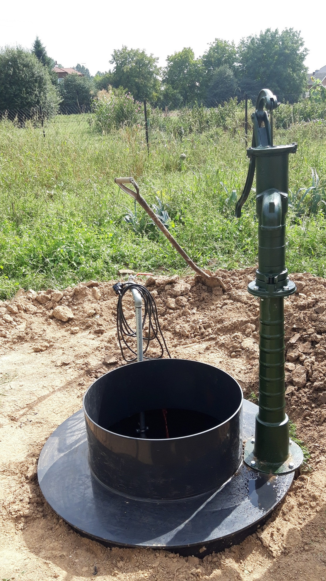 Zewnętrzna studnia z ręczną pompą w kolorze zielonym, otoczona ziemią i trawą. W tle widoczna łopata oparta o ziemię i zieleń.