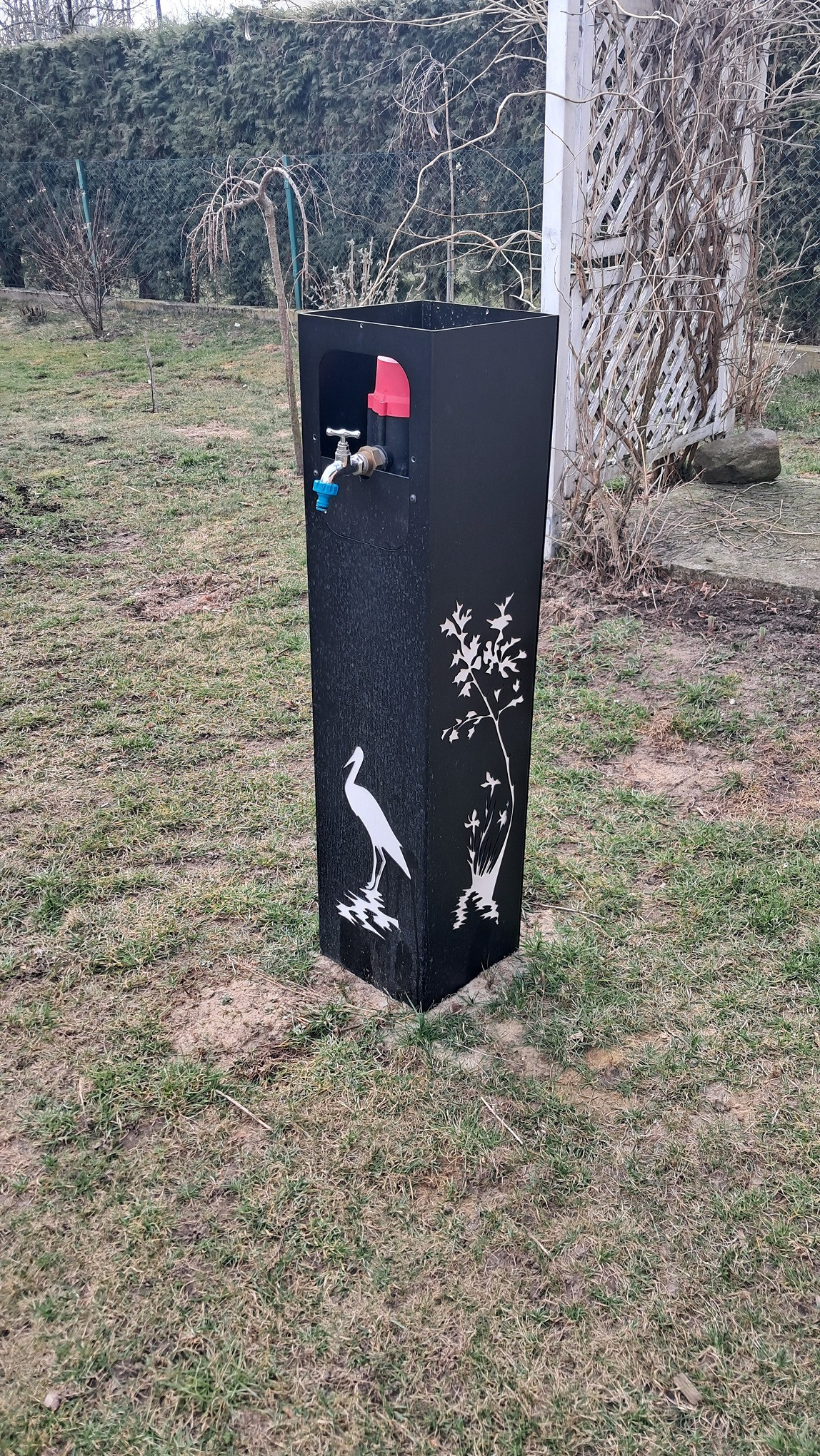 Czarna, dekoracyjna studzienka ogrodowa z kranem, zdobiona motywem czapli i drzewa, umieszczona na trawniku w ogrodzie. W tle widoczna pergola i drzewa.