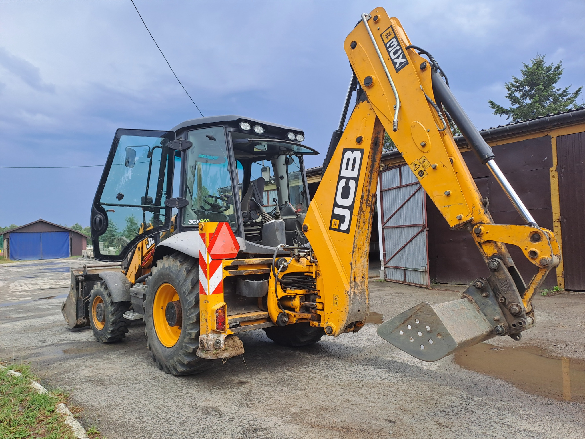 Żółto-szara koparko-ładowarka JCB 3CX z otwartą kabiną na betonowym placu, widok z tyłu, z łyżką skierowaną w dół. W tle budynki i pochmurne niebo.