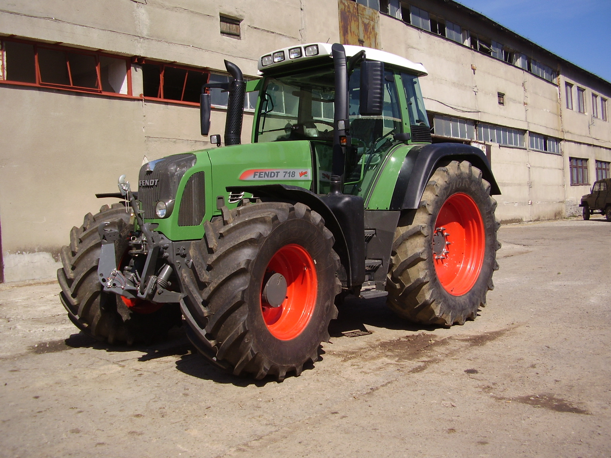 Zielony ciągnik Fendt 718 Vario z pomarańczowymi felgami na tle betonowego budynku z oknami. Widoczny fragment nieba i inny pojazd w oddali.