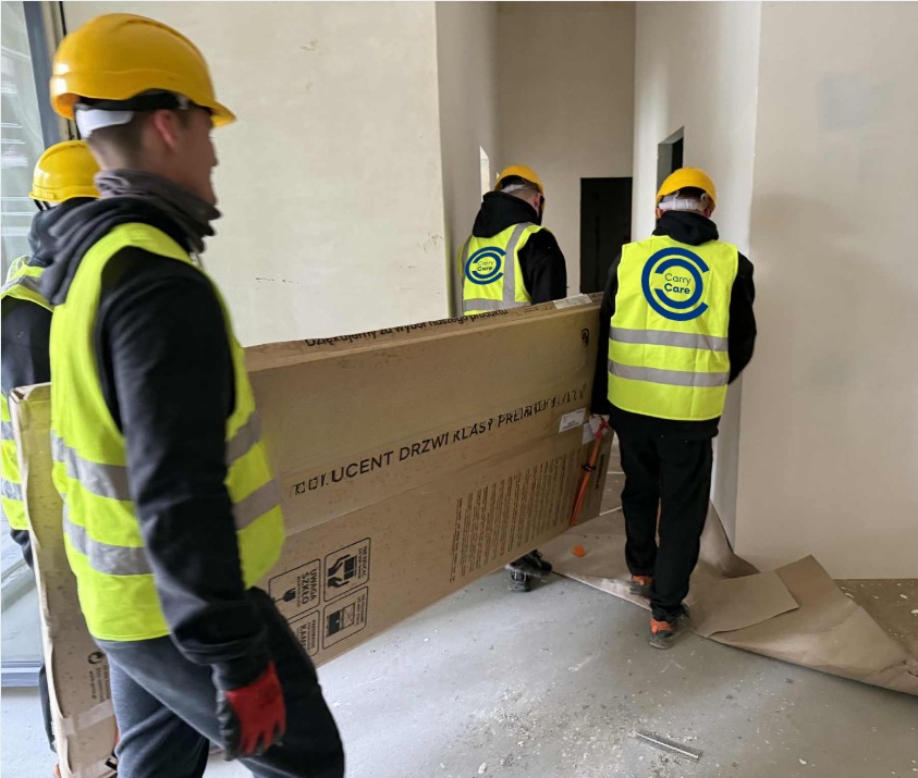 Ekipa w żółtych kamizelkach transportuje zapakowane drzwi w kartonie. Widoczne logo firmy transportowej na kamizelkach. Wnętrze w trakcie wykończenia.
