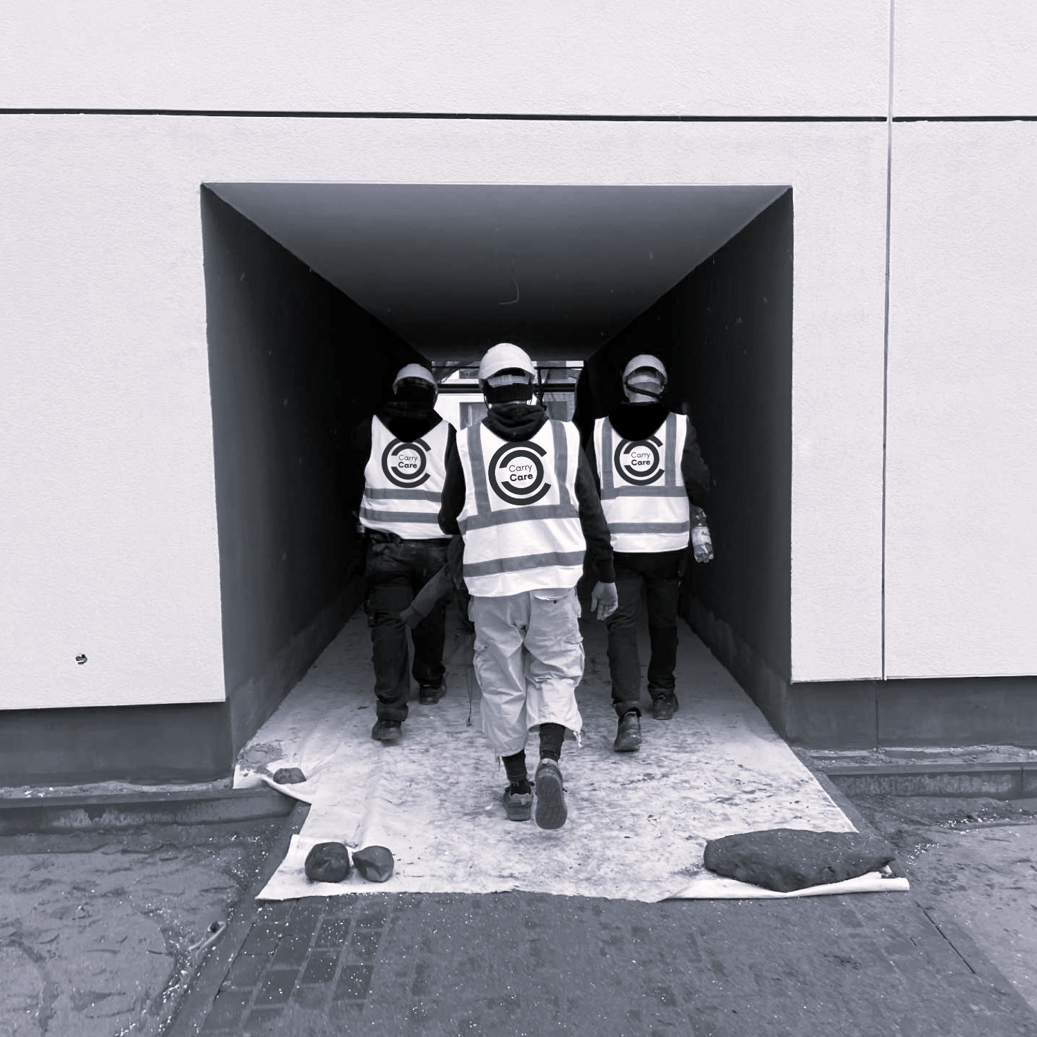 Trzech pracowników w kamizelkach odblaskowych wchodzi do tunelu. Jeden z nich ma kask. Podłoże zabezpieczone folią. Zdjęcie w czerni i bieli.