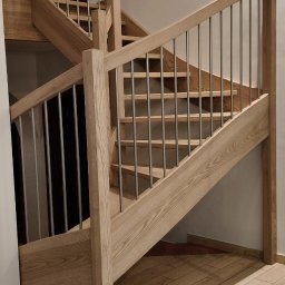 Jasne schody drewniane z metalową balustradą, widok z dołu, podkreślający naturalne usłojenie drewna i nowoczesny design. Ułożone płytki podłogowe imitujące drewno.