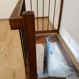 Fragment balustrady drewnianej z czarnymi, metalowymi prętami, zabezpieczającej schody prowadzące na antresolę. Widoczne drewniane stopnie i podłoga.