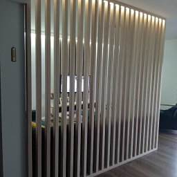 DREWLINE JAROSŁAW KISIELEWICZ - Ażurowa ścianka działowa z jasnego drewna, podświetlona od góry, oddzielająca przestrzeń w nowoczesnym wnętrzu. Widoczny fragment podłogi i wyposażenia pokoju.