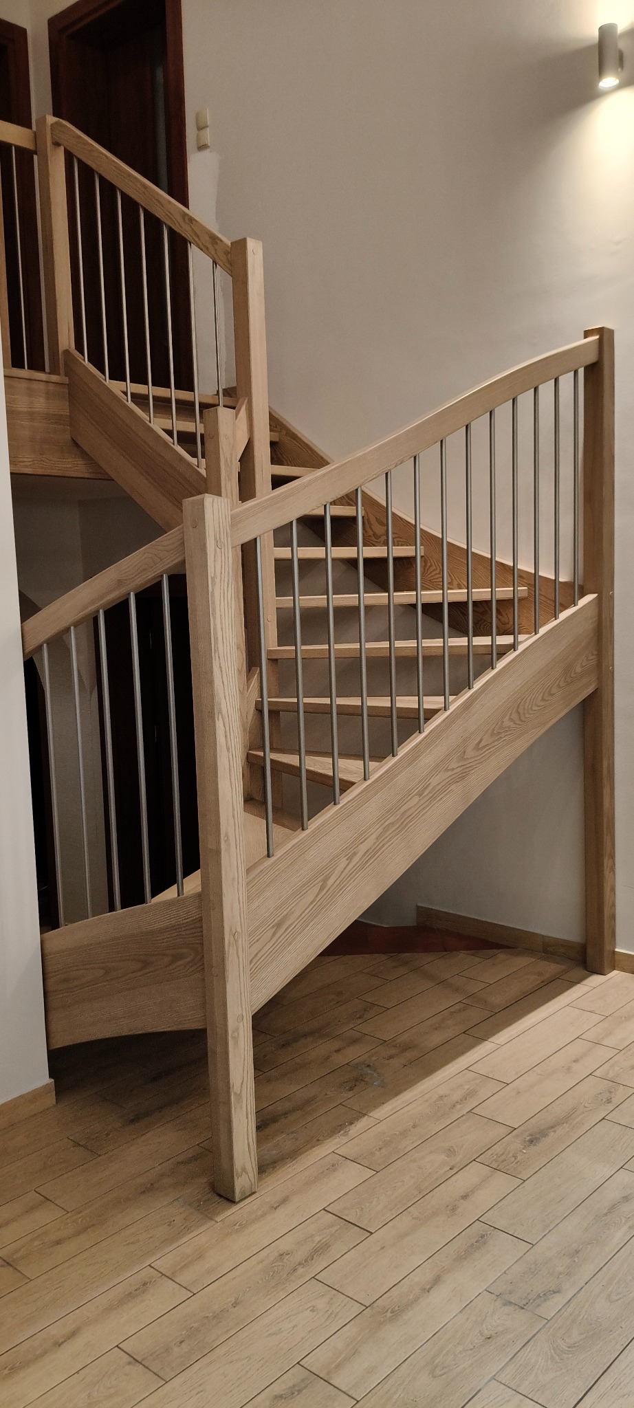 Jasne schody drewniane z metalową balustradą, widok z dołu, podkreślający naturalne usłojenie drewna i nowoczesny design. Ułożone płytki podłogowe imitujące drewno.