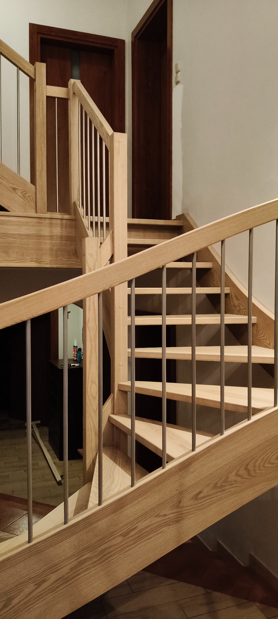 Jasne, drewniane schody z metalowymi balustradami w nowoczesnym wnętrzu. Widoczne drzwi w tle i fragment podłogi. Ujęcie z dołu, podkreślające konstrukcję schodów.