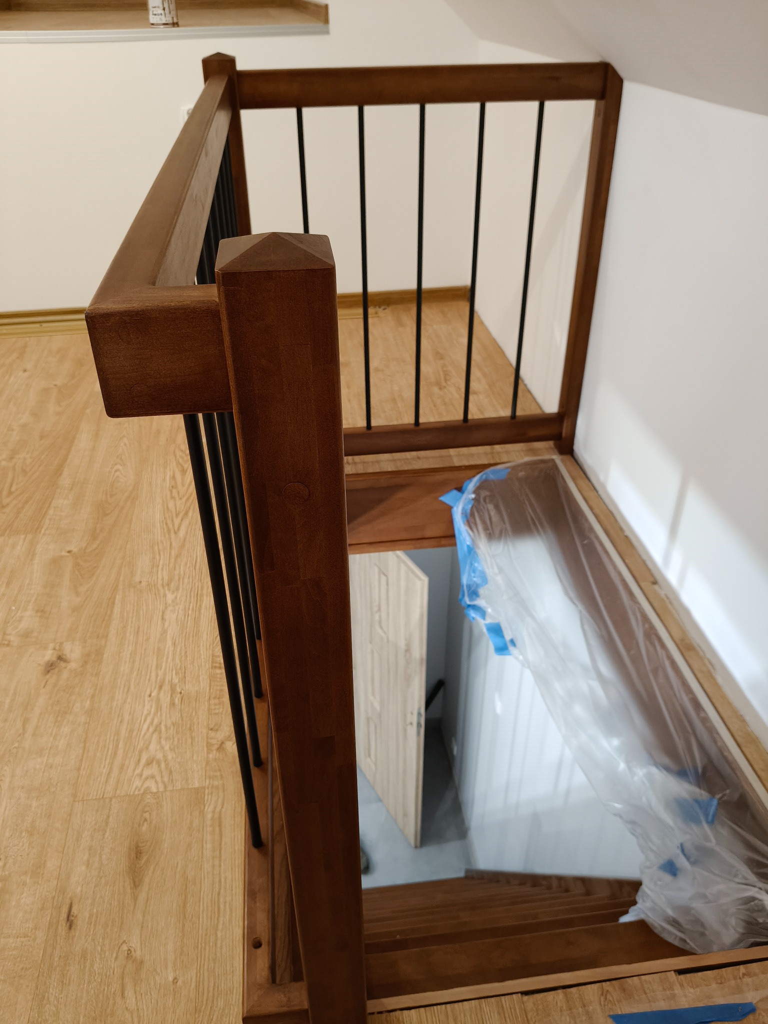 Fragment balustrady drewnianej z czarnymi, metalowymi prętami, zabezpieczającej schody prowadzące na antresolę. Widoczne drewniane stopnie i podłoga.
