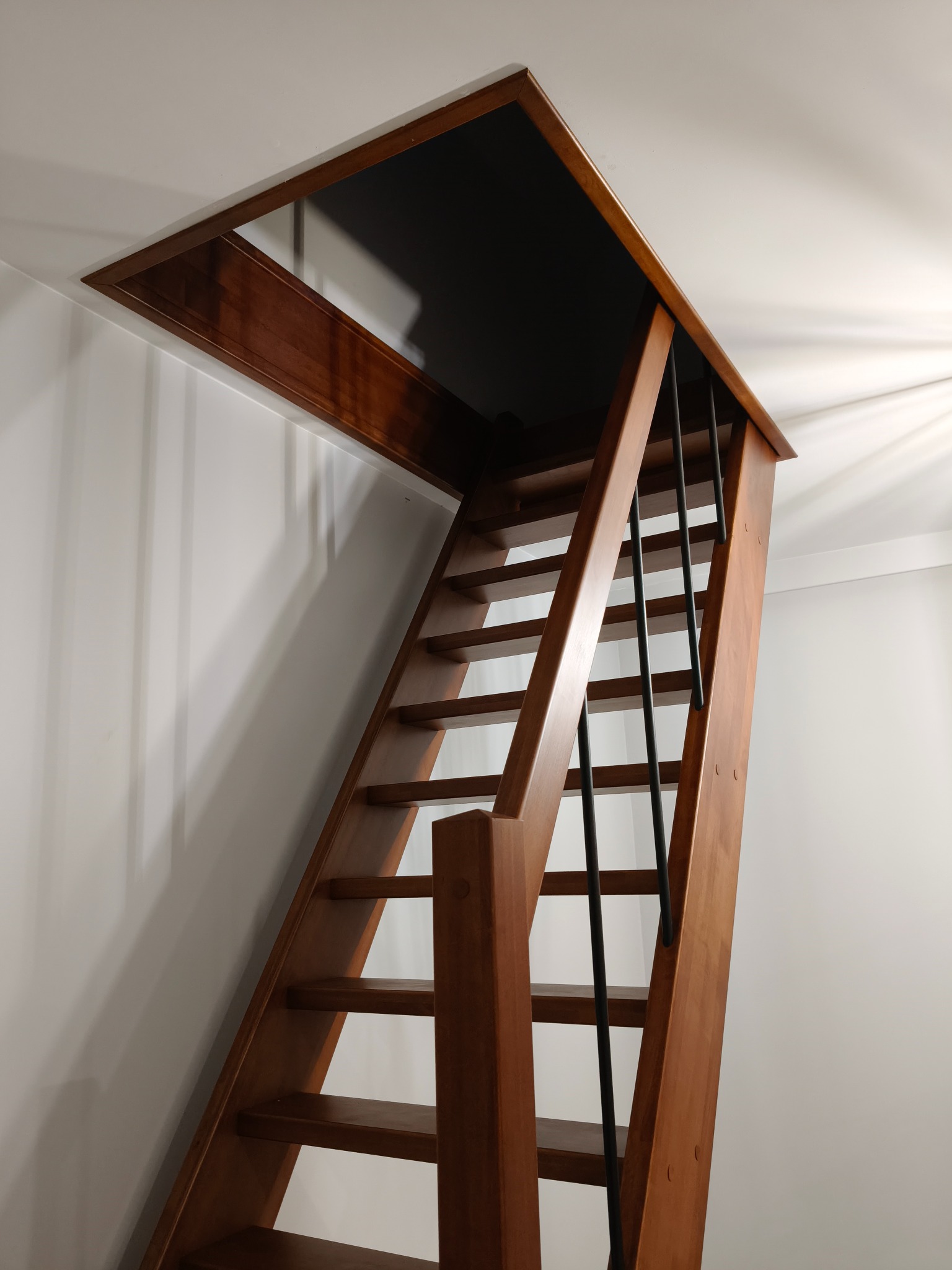 Drewniane schody strychowe z czarnymi, metalowymi balustradami, prowadzące do otworu w suficie. Ciepłe oświetlenie podkreśla naturalny odcień drewna.