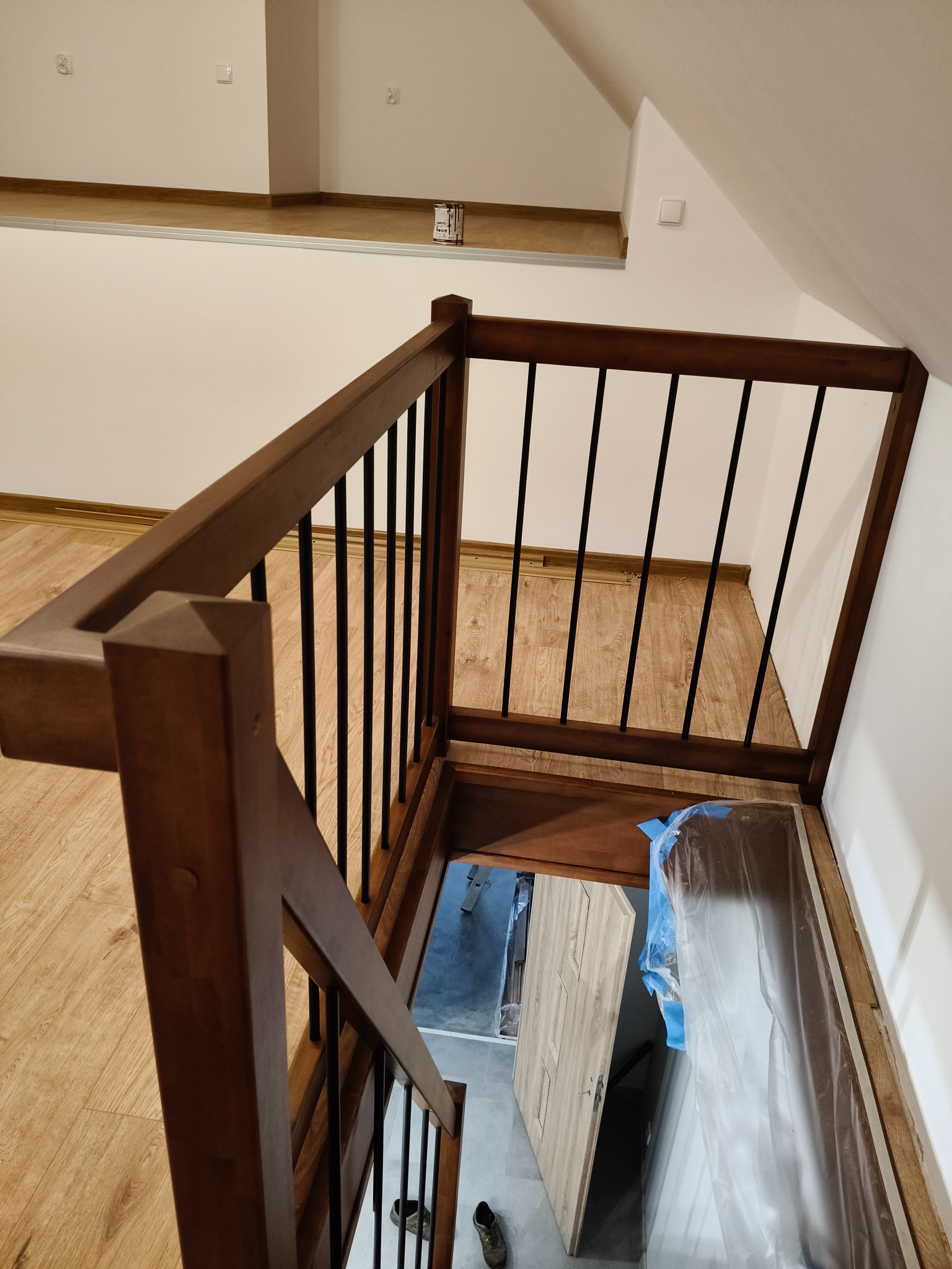 Drewniane schody z czarnymi, metalowymi balustradami, widziane z góry. Schody prowadzą do piwnicy, gdzie widać zabezpieczone folią drzwi i podłogę.