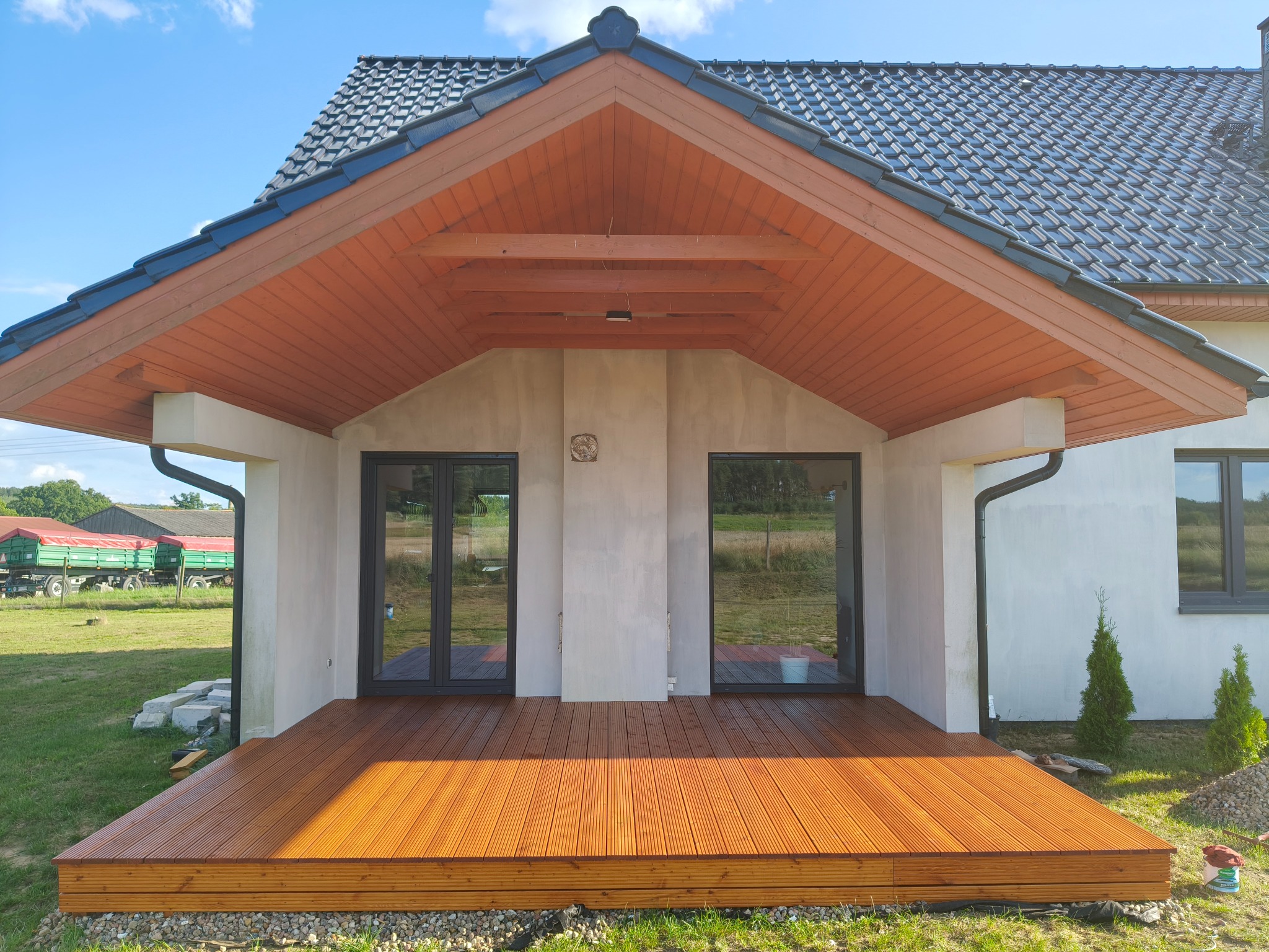 Zadaszony taras z desek w odcieniach brązu, z widokiem na ogród i budynek z szarą dachówką. Widoczne detale wykończenia i naturalne otoczenie.