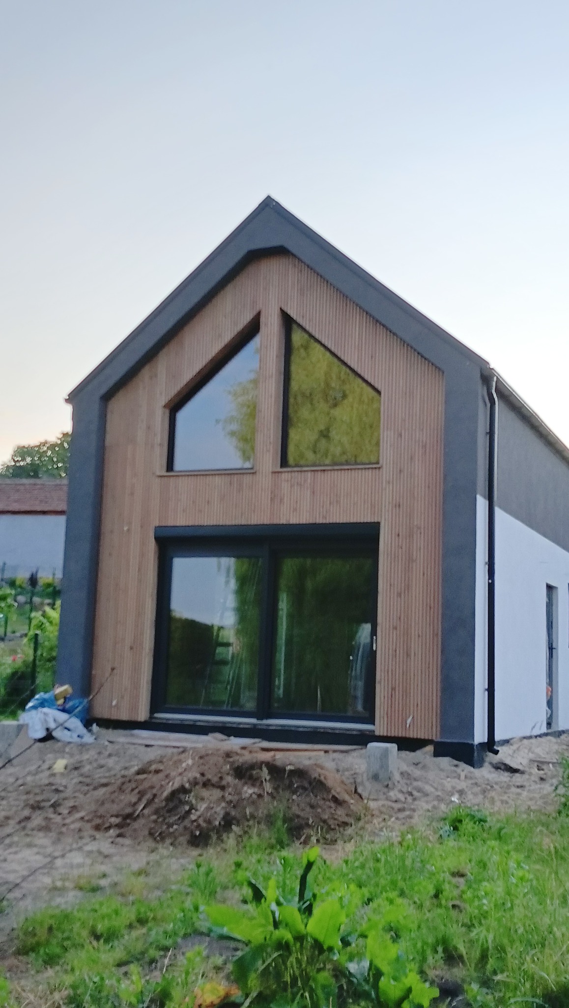 Nowoczesny dom z drewnianą elewacją i dużymi oknami, w trakcie wykończenia. Widoczne detale drewnianej fasady i otoczenie budynku.