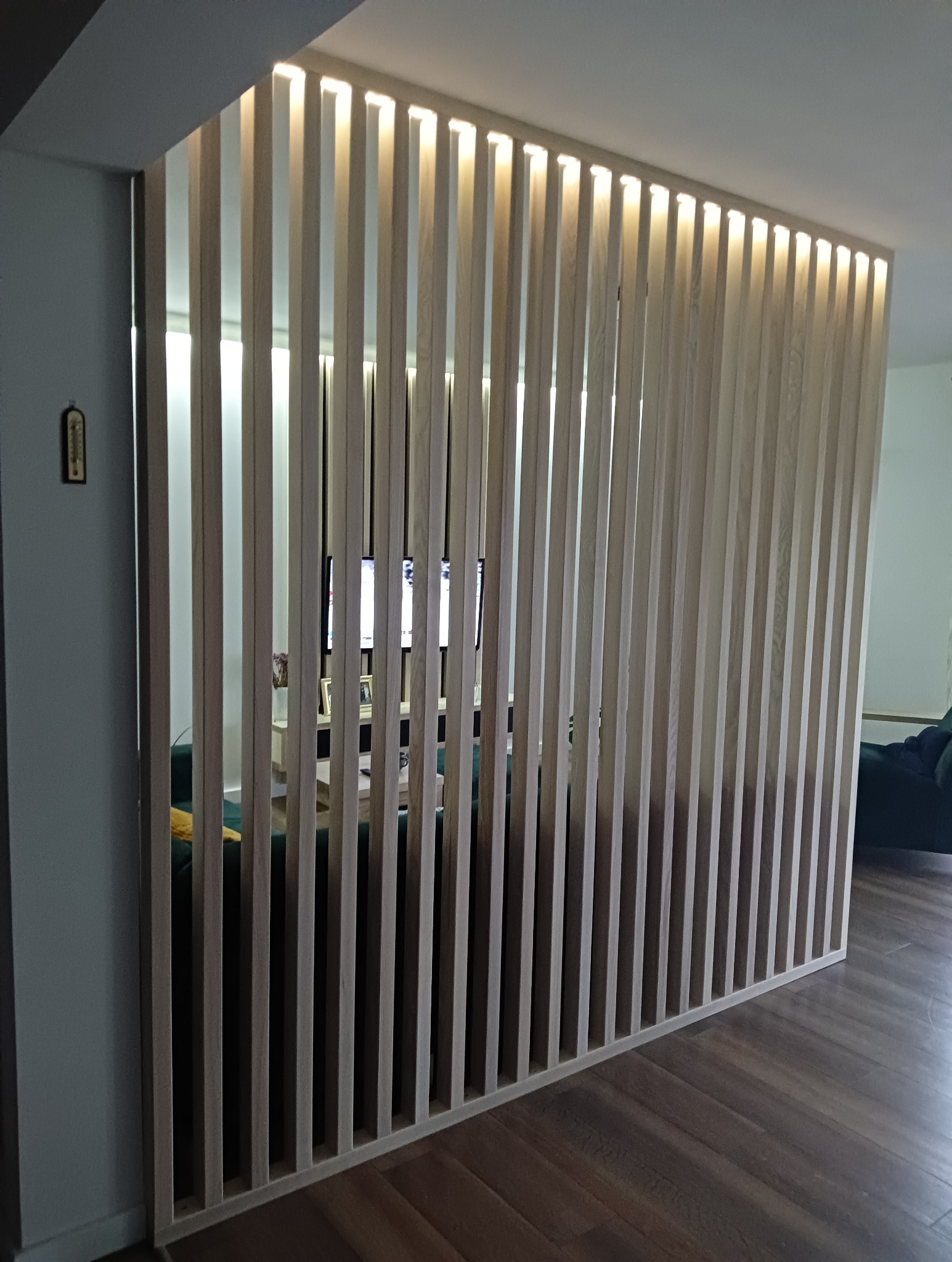 Ażurowa ścianka działowa z jasnego drewna, podświetlona od góry, oddzielająca przestrzeń w nowoczesnym wnętrzu. Widoczny fragment podłogi i wyposażenia pokoju.