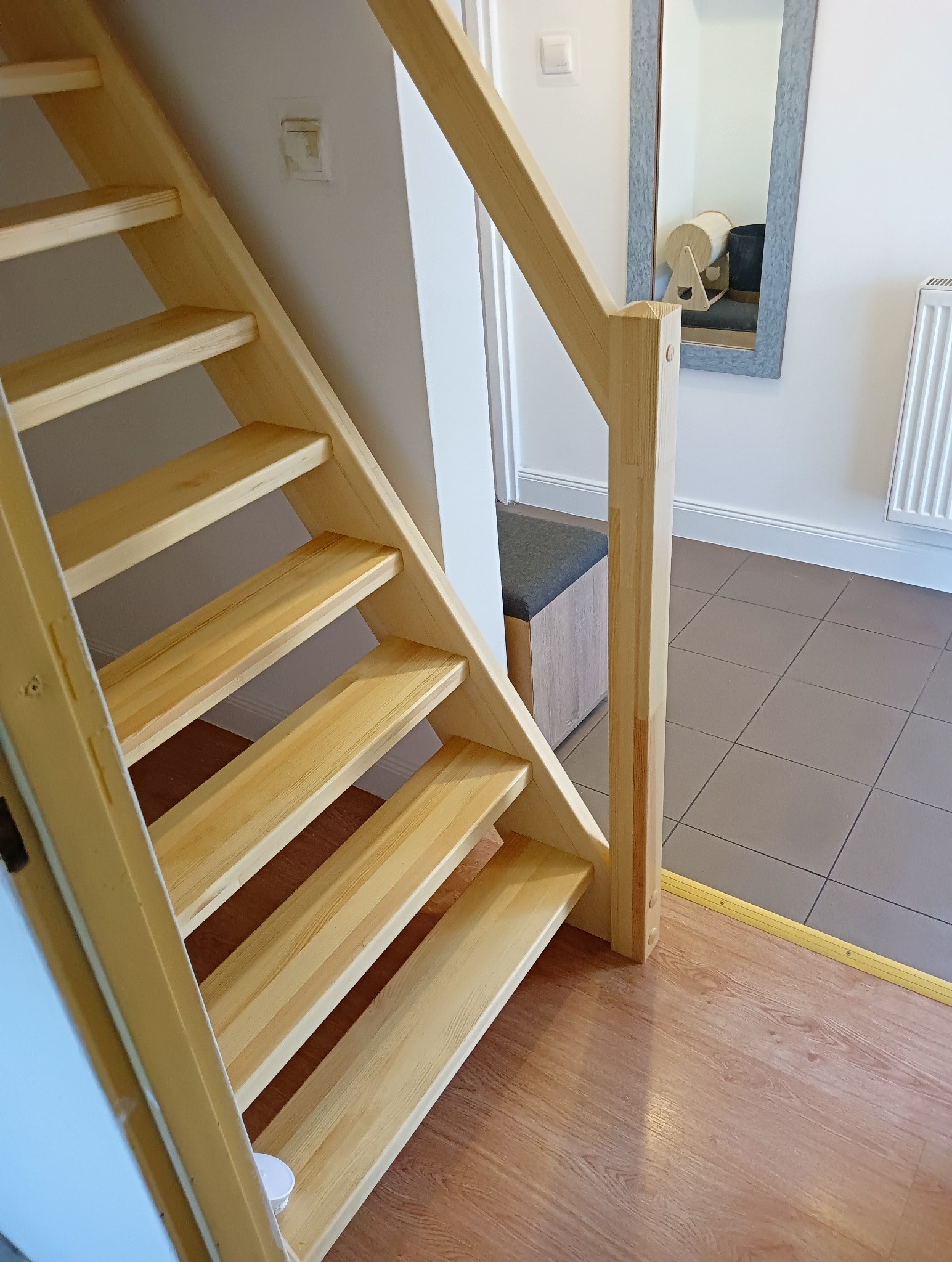 Jasne, proste schody z drewna sosnowego, prowadzące do góry. Widoczne stopnie, barierka i fragment podłogi. W tle lustro i grzejnik.