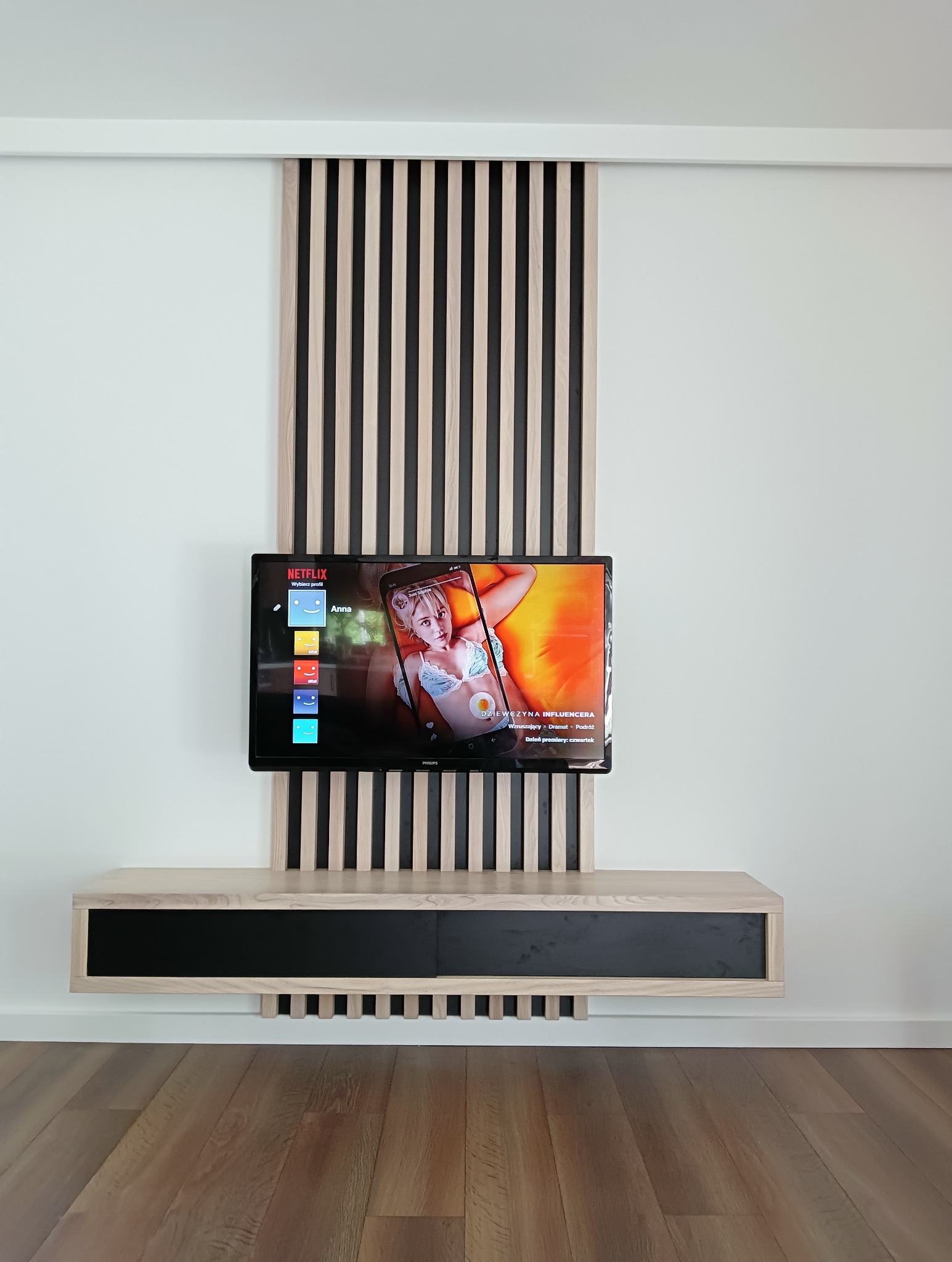 Nowoczesna ściana TV z drewnianymi lamelami w odcieniach beżu i czerni, podwieszana szafka RTV z czarnym frontem, drewniana podłoga. Minimalistyczny design.