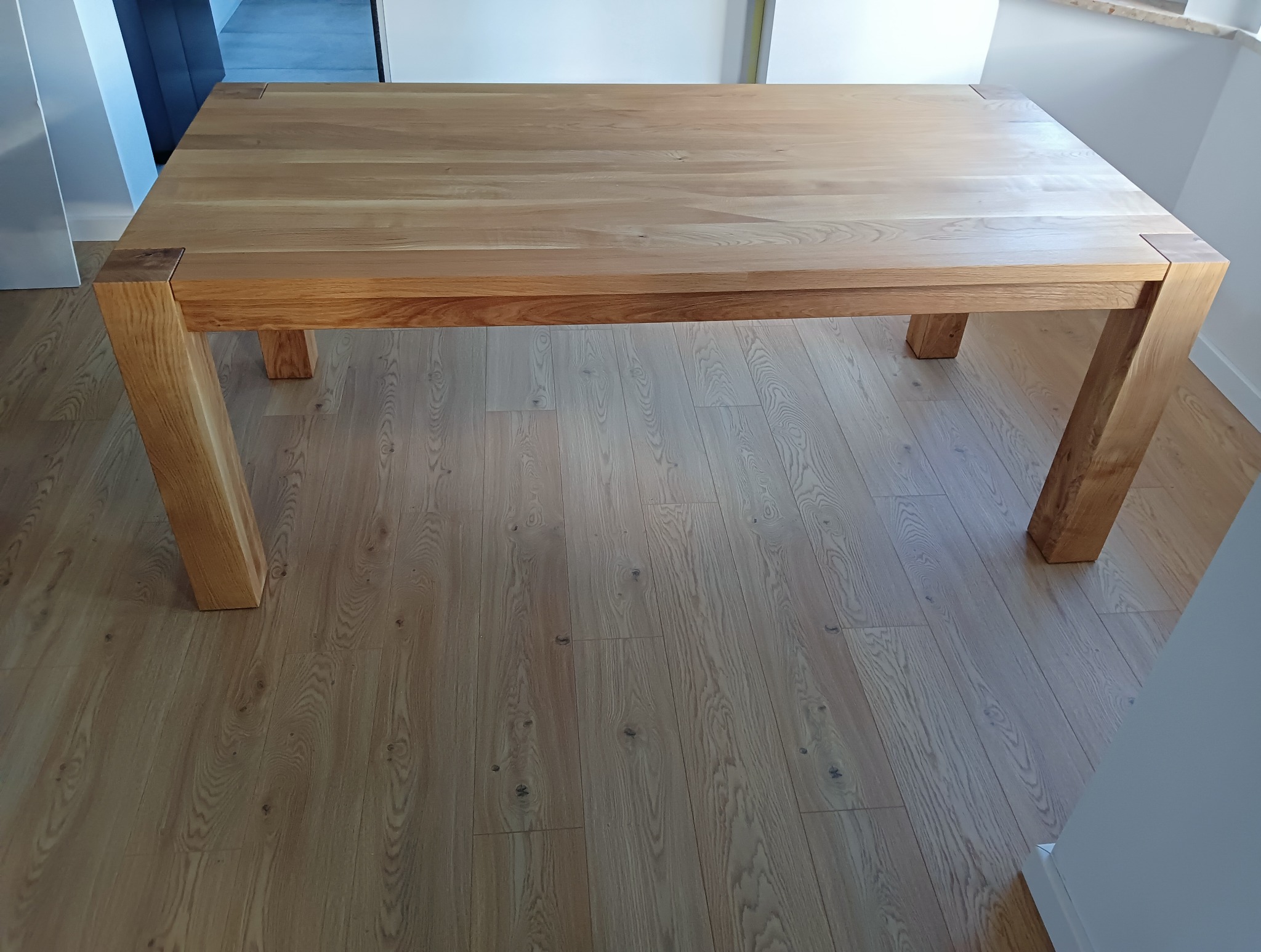 Solidny, jasny stół z drewna dębowego z grubym blatem i nogami, widoczny na tle drewnianej podłogi. Minimalistyczny design, naturalne wykończenie.