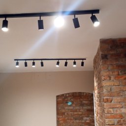 Mariusz Staniszewski - Nowoczesne oświetlenie sufitowe na szynoprzewodach w biurze z ceglaną ścianą. Widoczne biurka i krzesła biurowe. Minimalistyczny design.