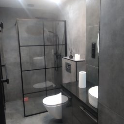 Mariusz Staniszewski - Nowoczesna łazienka w odcieniach szarości z kabiną prysznicową typu walk-in z czarnymi profilami, wiszącą toaletą i szafką z umywalką nablatową.