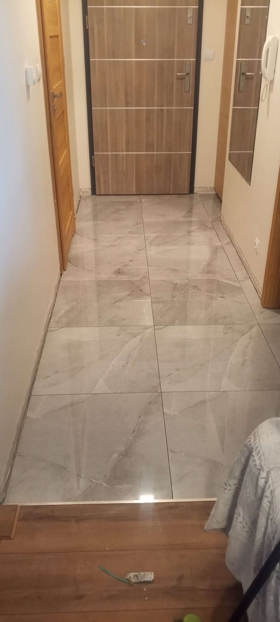 Jasne, marmurowe płytki podłogowe w korytarzu, z widocznymi drzwiami wejściowymi i fragmentem drewnianej podłogi na pierwszym planie. Widoczne fugi i listwy przypodłogowe.