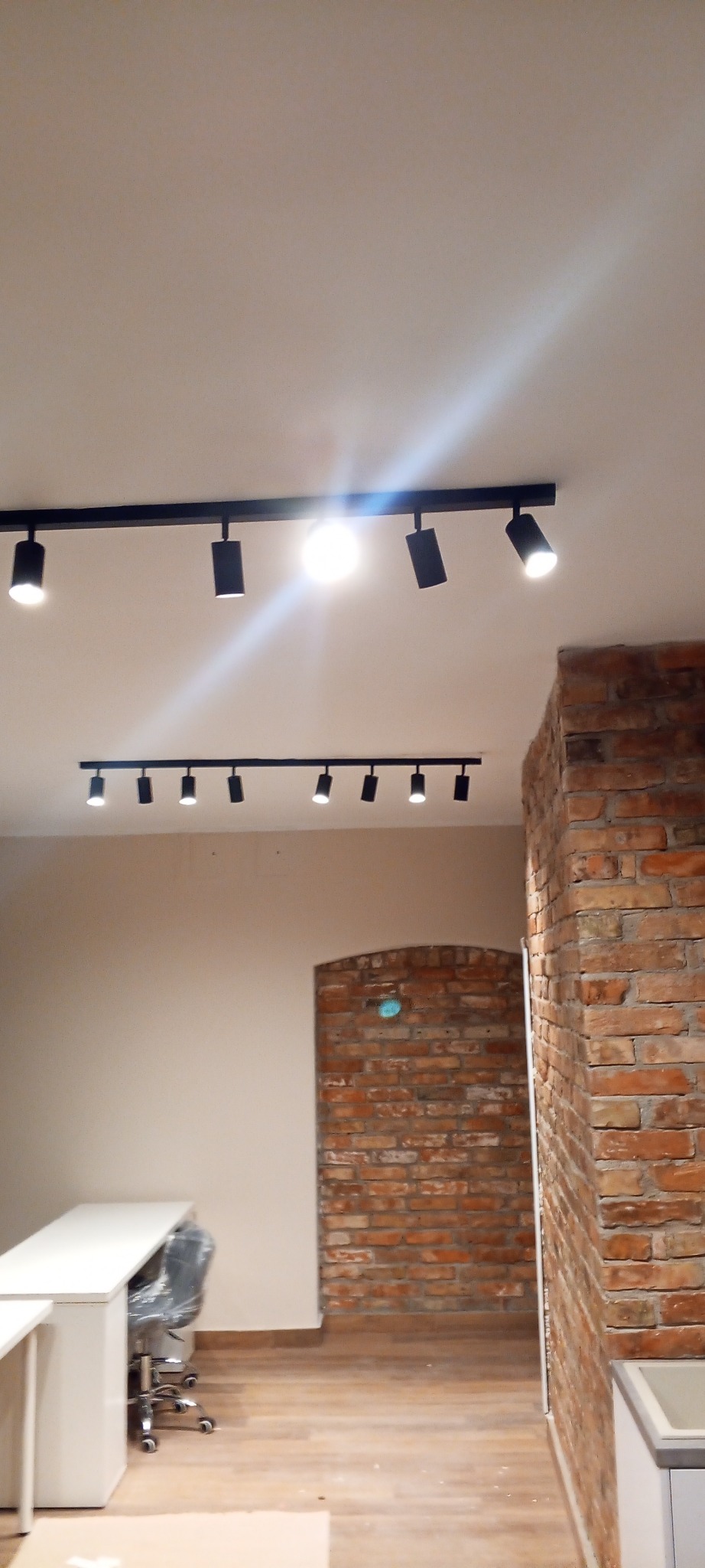 Nowoczesne oświetlenie sufitowe na szynoprzewodach w biurze z ceglaną ścianą. Widoczne biurka i krzesła biurowe. Minimalistyczny design.