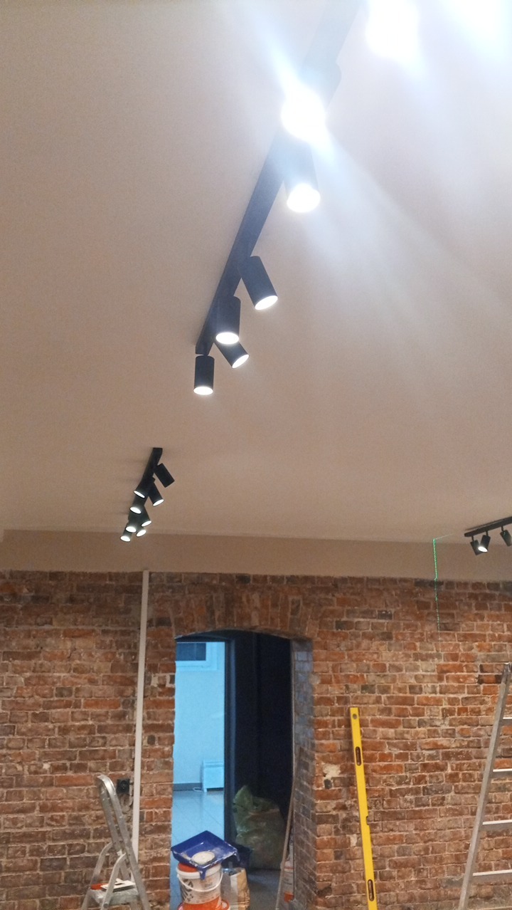 Czarne reflektory szynowe na suficie w pomieszczeniu z ceglaną ścianą i widocznymi narzędziami, sugerujące prace wykończeniowe.