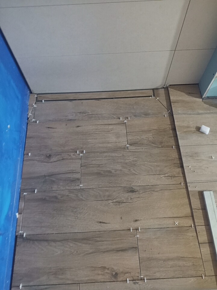 Układanie podłogi z płytek imitujących drewno, widoczne krzyżyki dystansowe, niebieska hydroizolacja na ścianie, etap prac wykończeniowych w łazience.