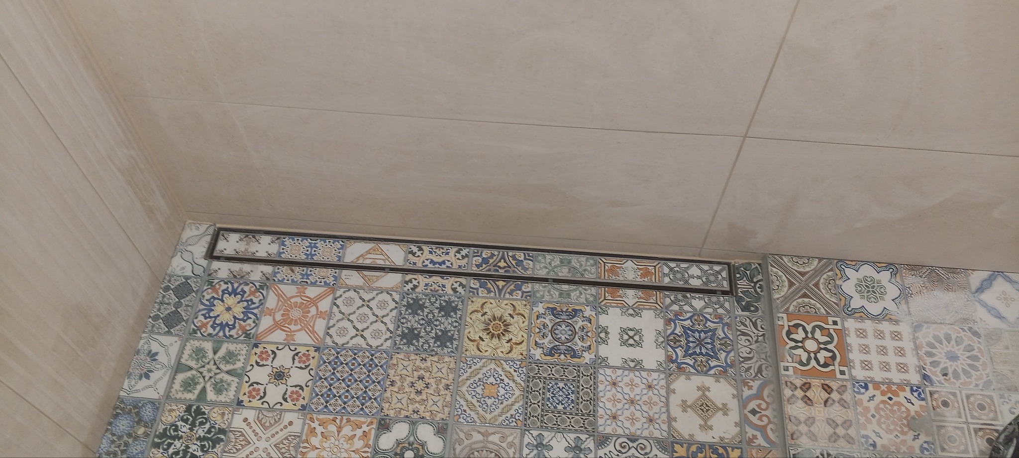 Wykończona łazienka z dekoracyjnymi, wzorzystymi płytkami ceramicznymi ułożonymi dookoła odpływu liniowego. Beżowe płytki na ścianach i suficie.