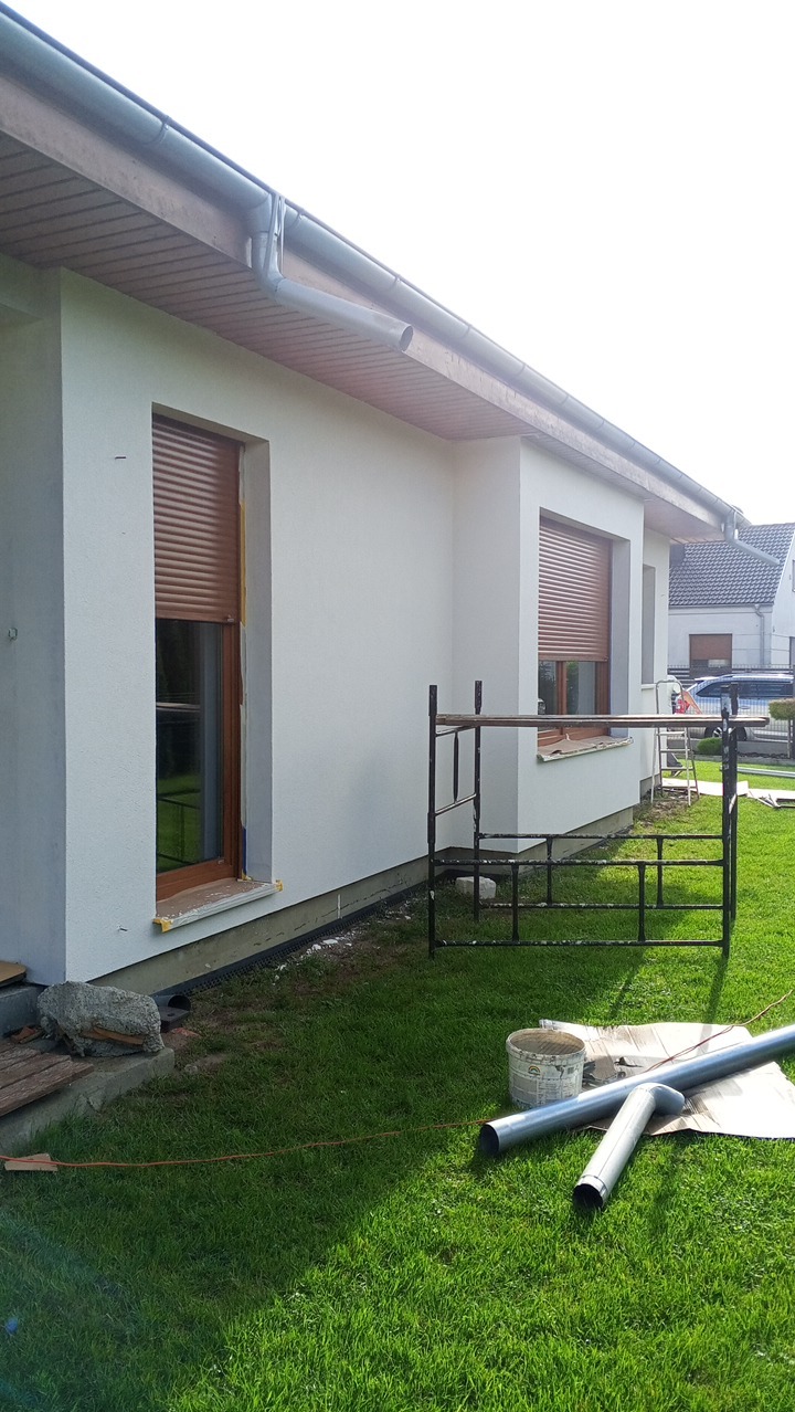 Biała elewacja domu jednorodzinnego z brązowymi roletami okiennymi. Widoczne rusztowanie, rynny i elementy instalacji na trawniku. Ujęcie z poziomu trawy.