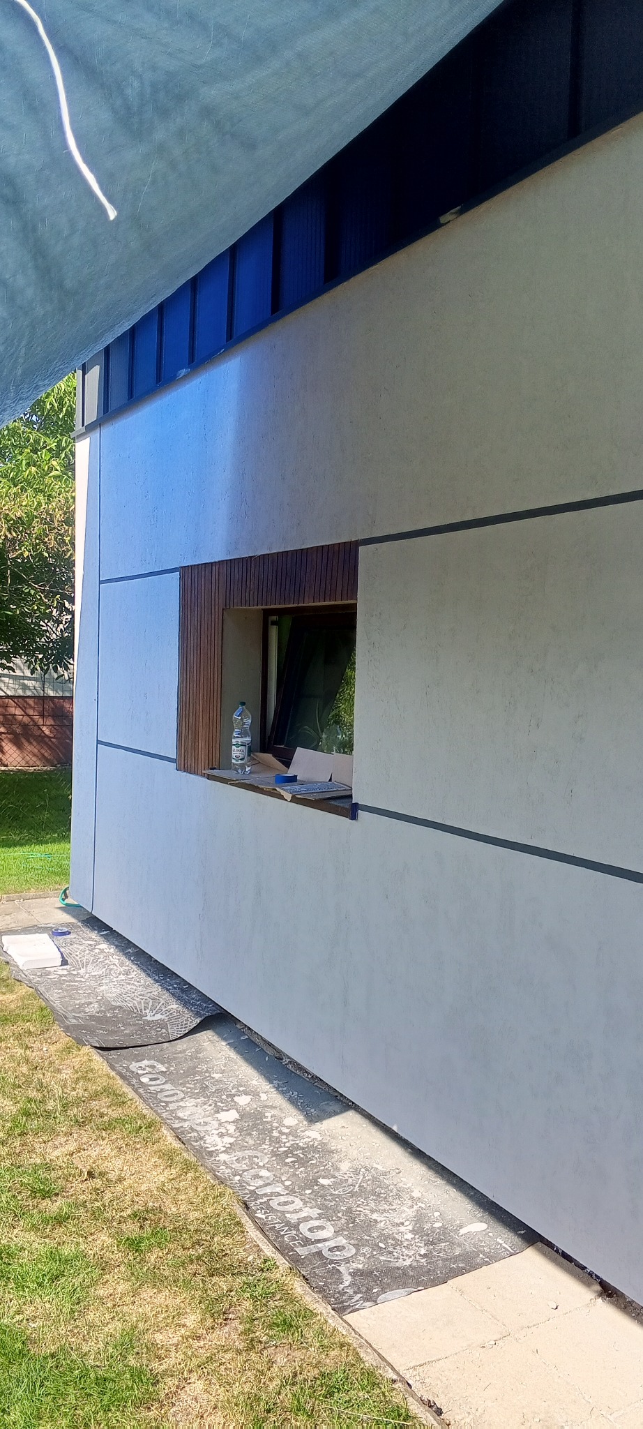 Elewacja budynku z oknem w drewnianej obudowie, widoczne fragmenty ocieplenia i zabezpieczenia podłoża. Świeżo położony tynk strukturalny.