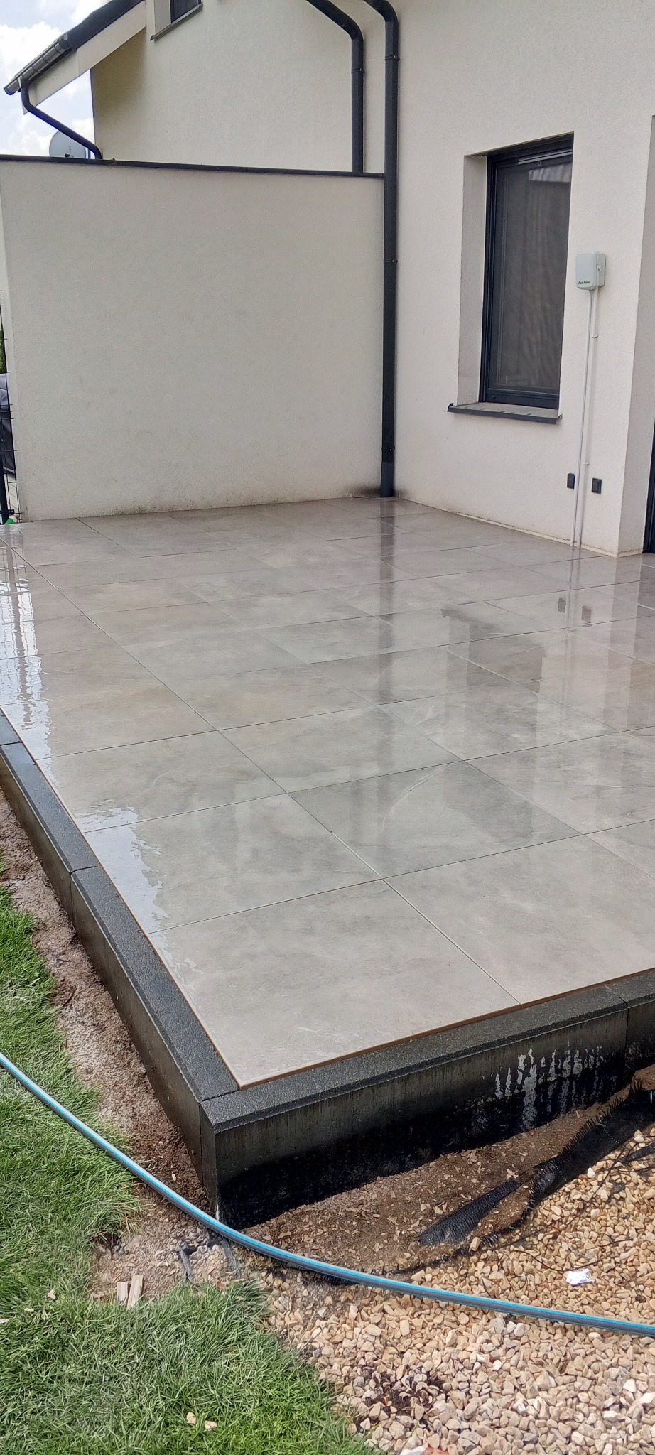 Wyłożony jasnymi płytkami taras z ciemną obwódką, przylegający do budynku z oknem. Na płytkach widoczne refleksy, sugerujące świeżo umytą powierzchnię.