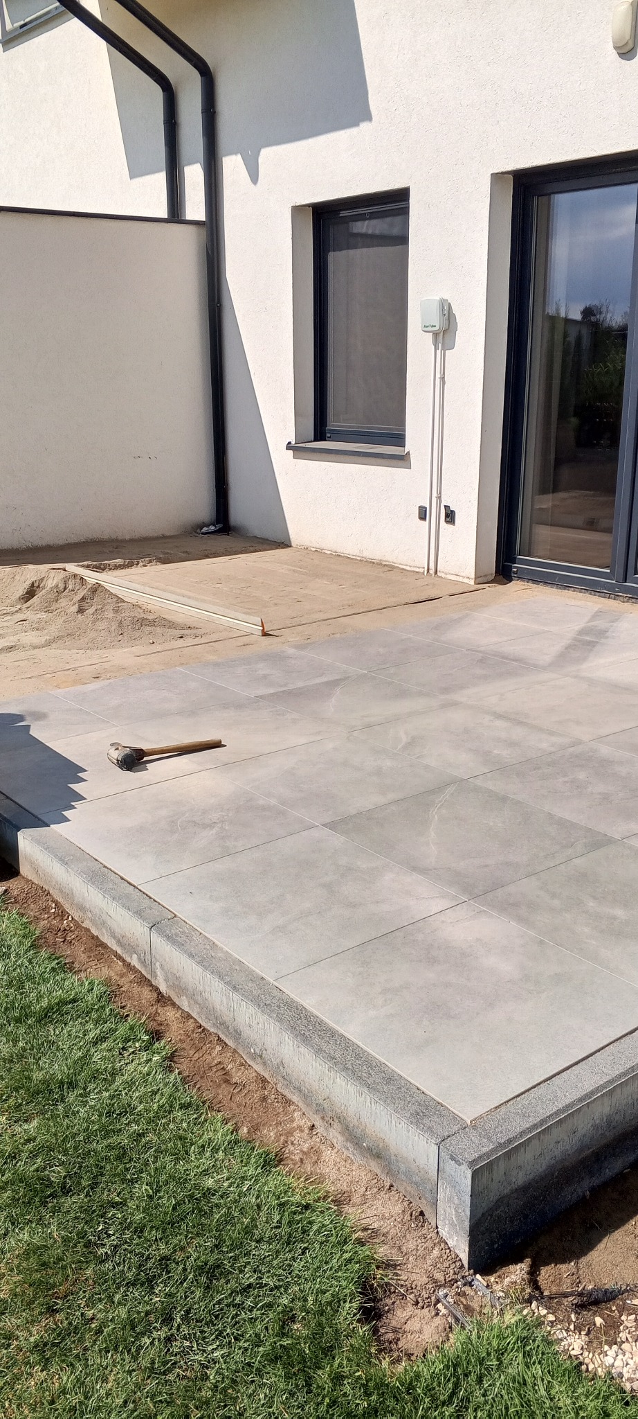 Jasnoszara, kwadratowa nawierzchnia tarasu z obrzeżem, częściowo ułożona przy białym budynku. Gumowy młotek leży na płytach. W tle piasek i przygotowany teren.