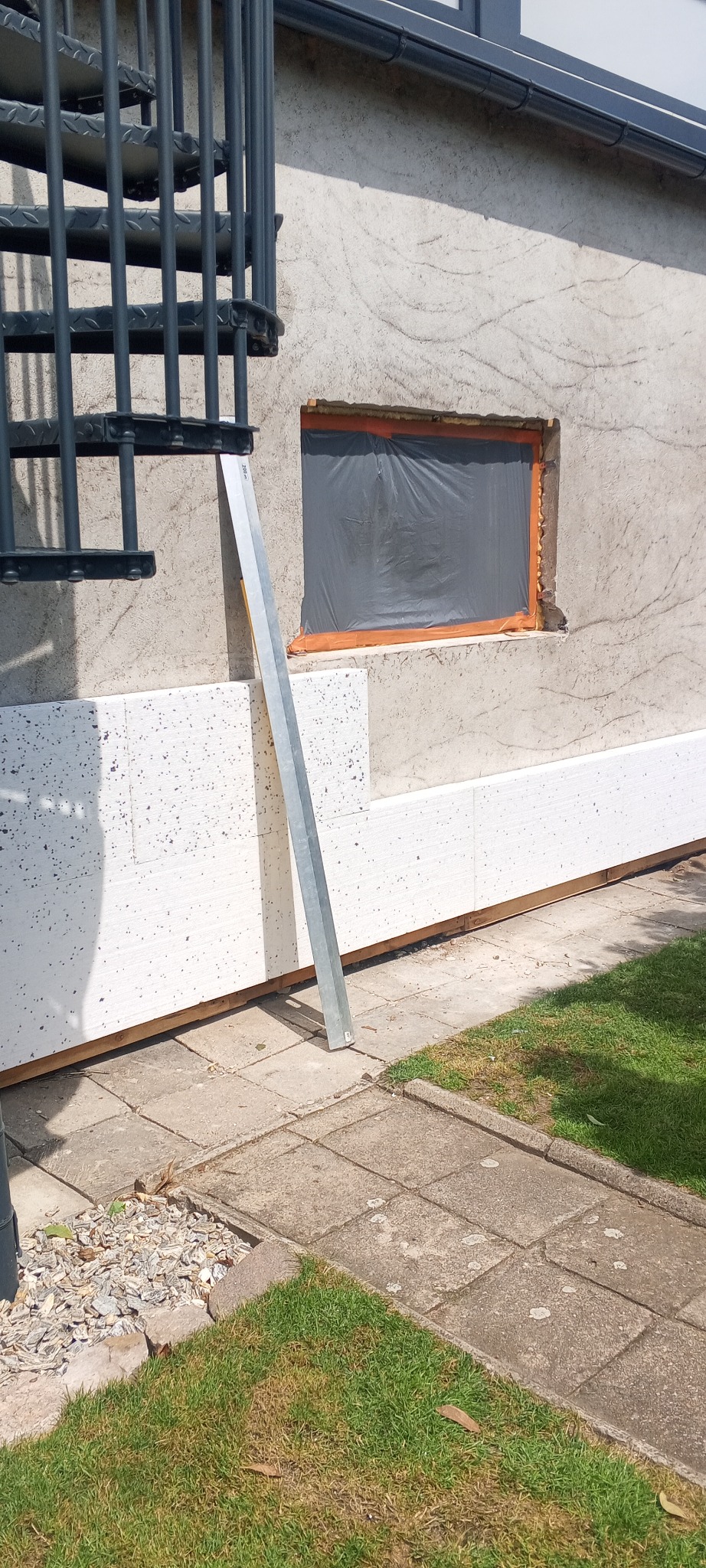 Ocieplenie ściany zewnętrznej budynku z widoczną warstwą styropianu i zabezpieczonym oknem folią. Metalowa listwa oparta o ścianę. Fragment schodów spiralnych.