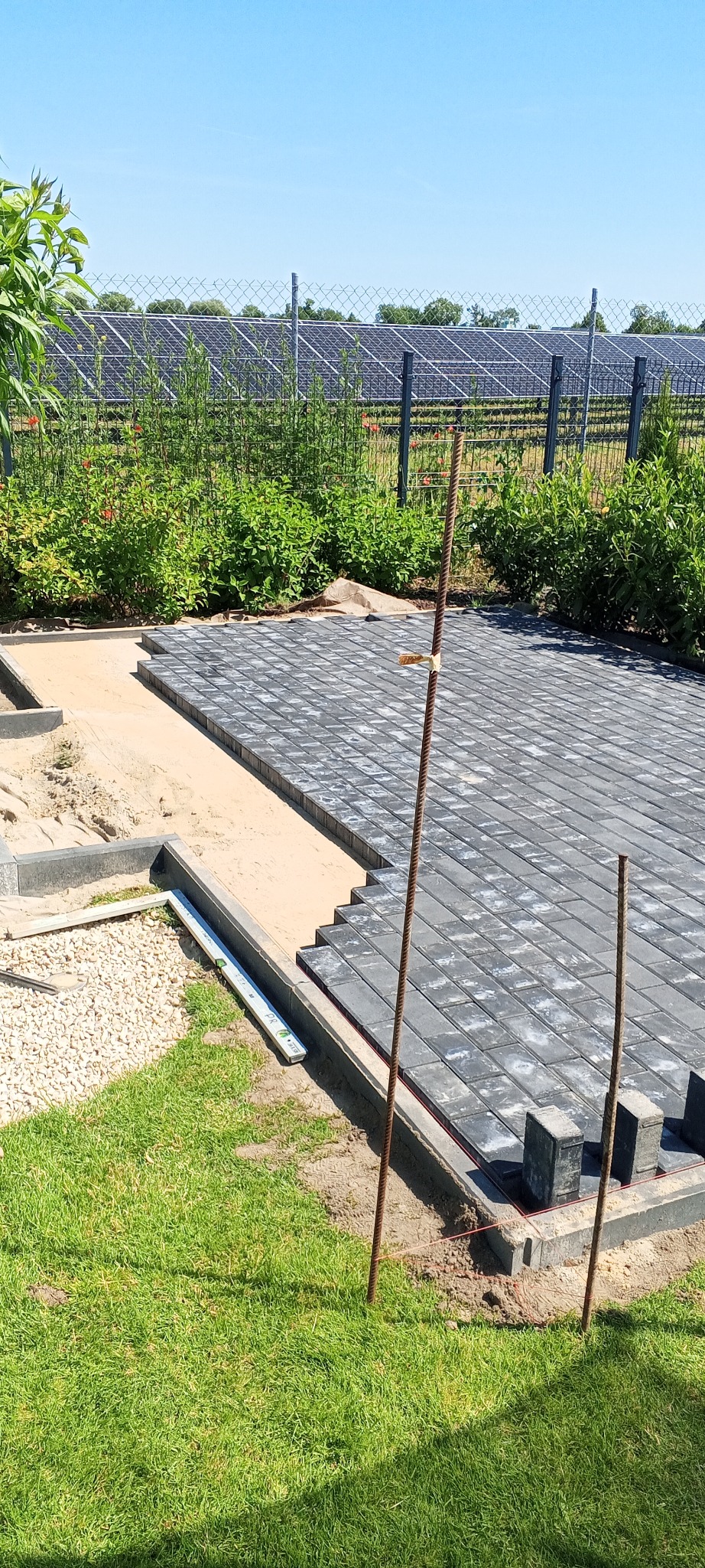 Układanie szarej kostki brukowej na podsypce piaskowej, widoczne narzędzia i poziomica, w tle ogrodzenie z siatki i panele słoneczne. Prace brukarskie w toku.