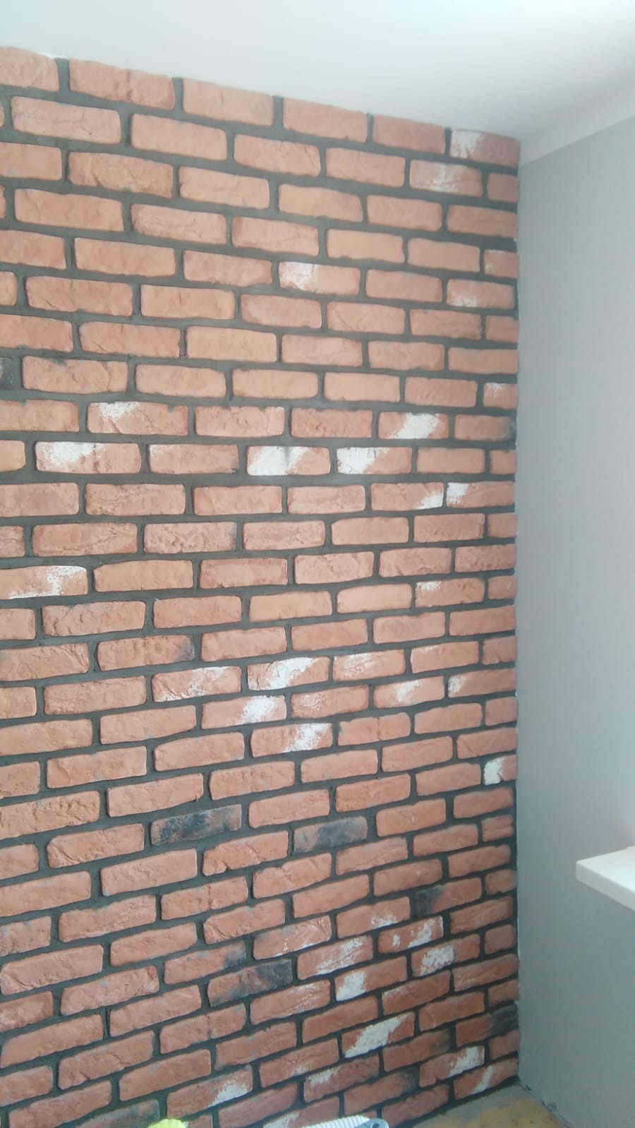 Ściana wykończona dekoracyjną cegłą w odcieniach czerwieni i brązu, z fugami w kolorze grafitowym. Fragment ściany pomalowany na szaro. Widok z dołu.