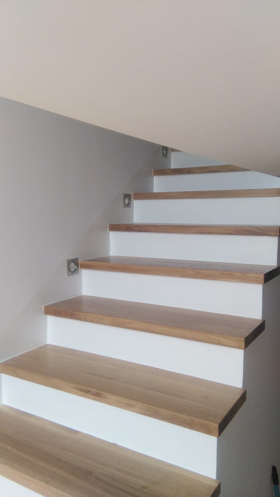 Nowoczesne schody z drewnianymi stopniami i białymi podstopnicami, oświetlone punktowymi lampkami w ścianie, widziane z dołu pod kątem.