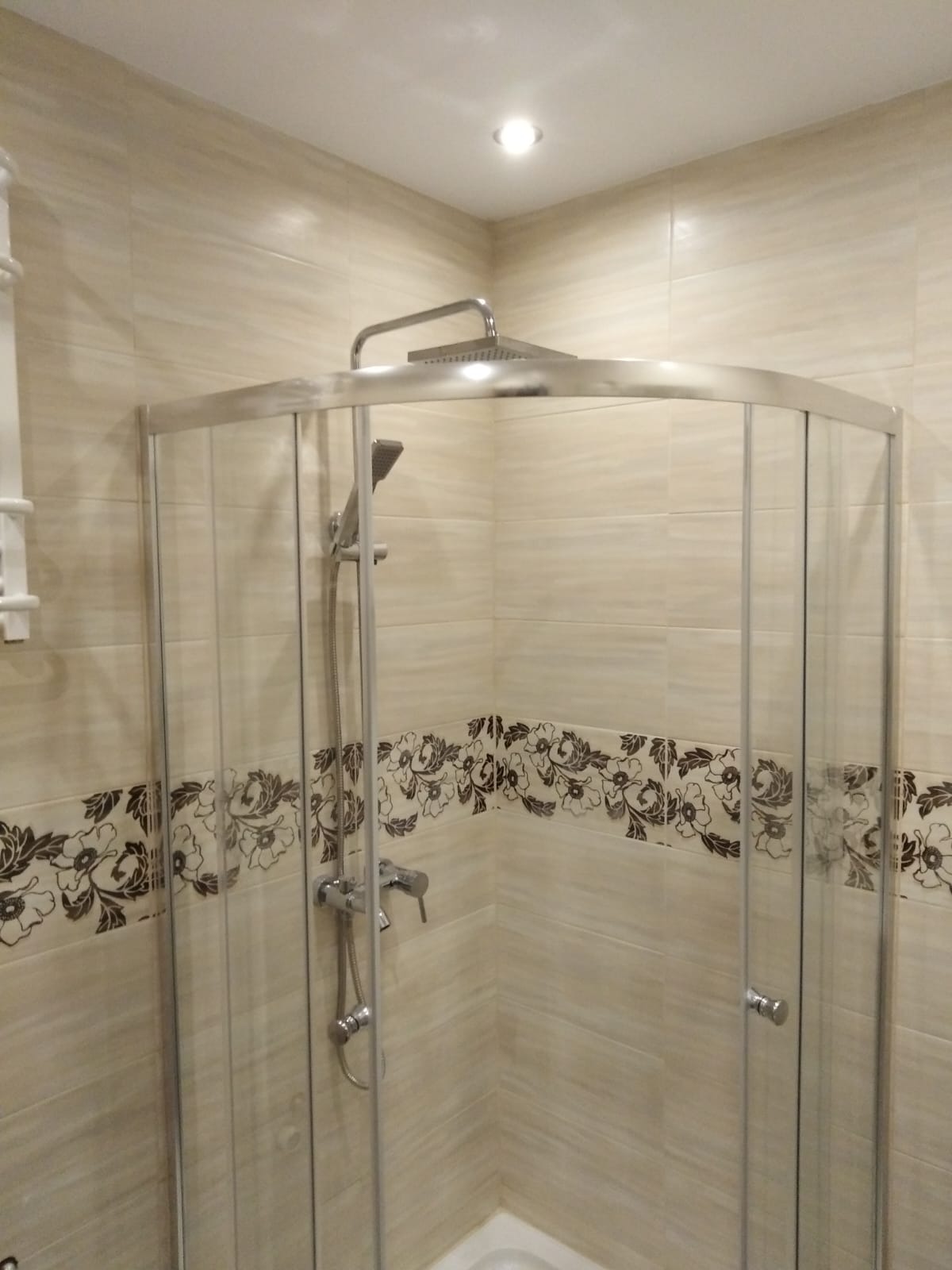 Kabina prysznicowa z beżowymi płytkami i dekoracyjnym pasem z motywem kwiatowym w łazience we Wrześni. Chromowana armatura i szklane drzwi.