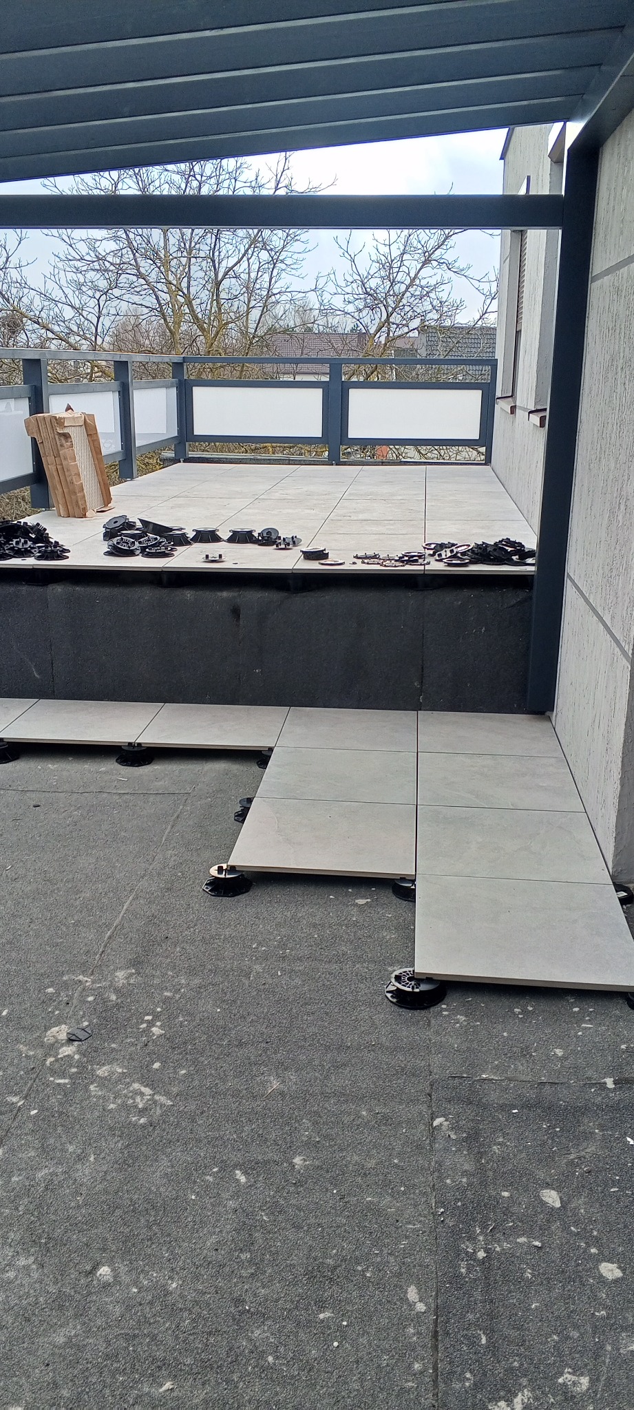 Montaż płytek na tarasie z użyciem regulowanych wsporników. Częściowo ułożone płytki w jasnym odcieniu szarości. Widoczne narzędzia i materiały montażowe.