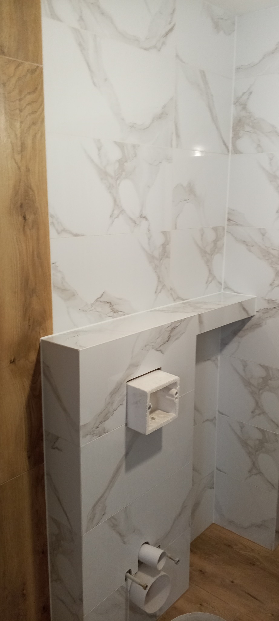 Wykończona łazienka z płytkami imitującymi marmur, zabudowa WC z półką. Widoczne przyłącza kanalizacyjne i fragment drewnopodobnej okładziny ściennej.