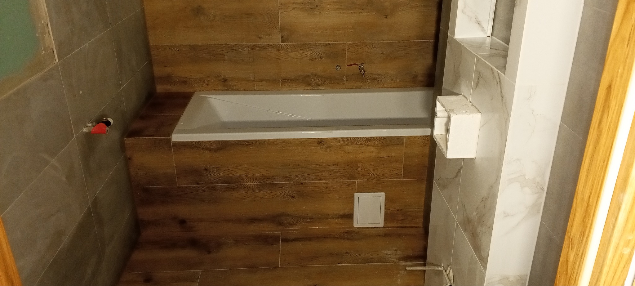 Biała wanna obudowana płytkami imitującymi drewno w łazience. Widoczne szare płytki na ścianach i białe elementy wykończeniowe. Ujęcie z góry.