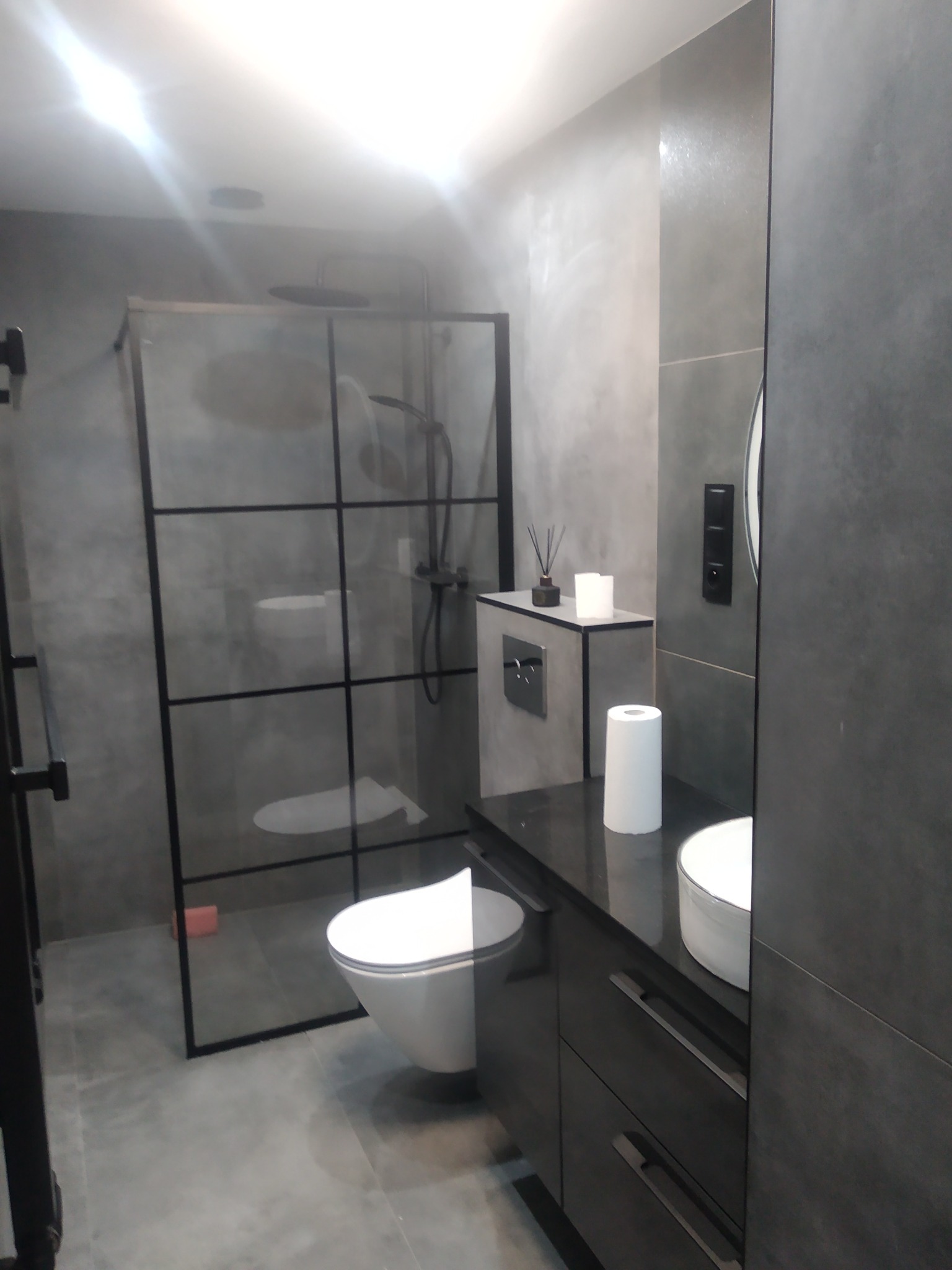 Nowoczesna łazienka w odcieniach szarości z kabiną prysznicową typu walk-in z czarnymi profilami, wiszącą toaletą i szafką z umywalką nablatową.
