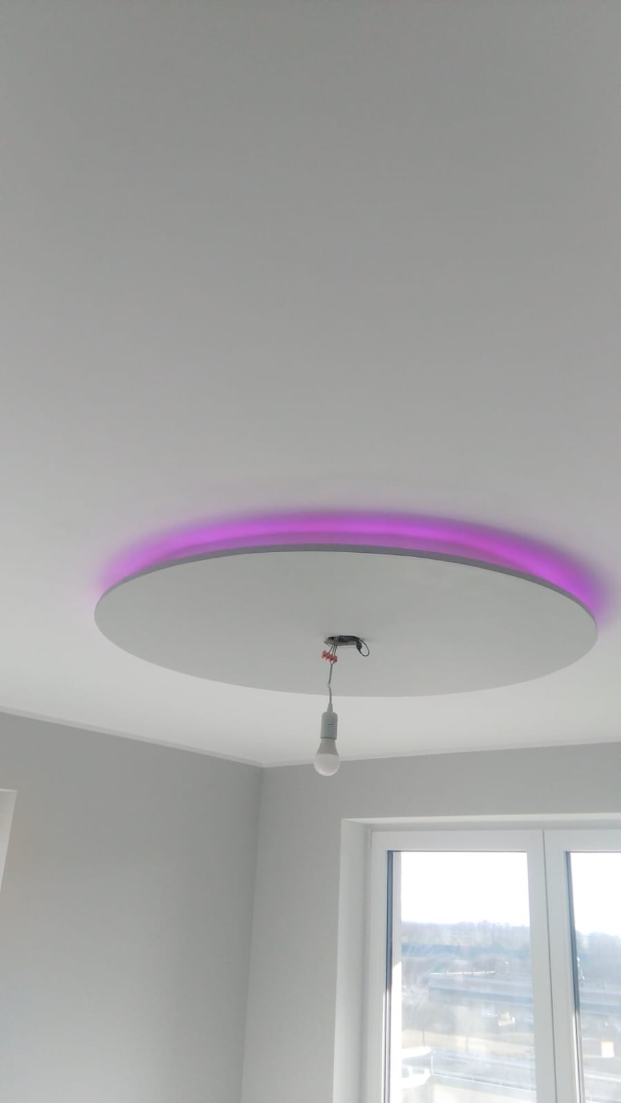 Okrągła, podwieszana konstrukcja sufitowa z fioletowym podświetleniem LED, widoczna w rogu pokoju z oknem. W tle szare ściany i sufit.