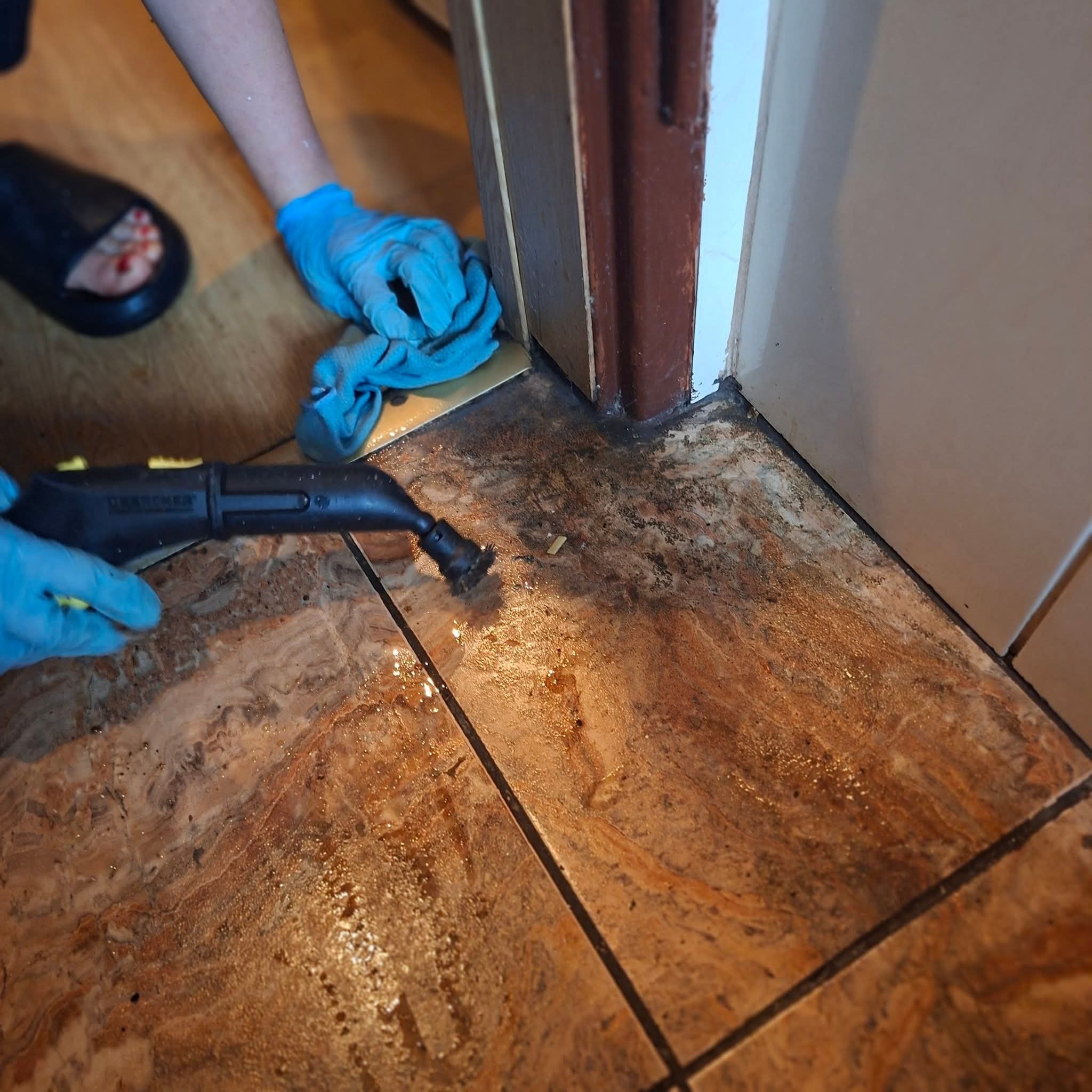 Czyszczenie parowe brudnej, brązowej podłogi z płytek przy listwie przypodłogowej. Użycie parownicy Karcher i niebieskiej ściereczki. Osoba w niebieskich rękawicach.
