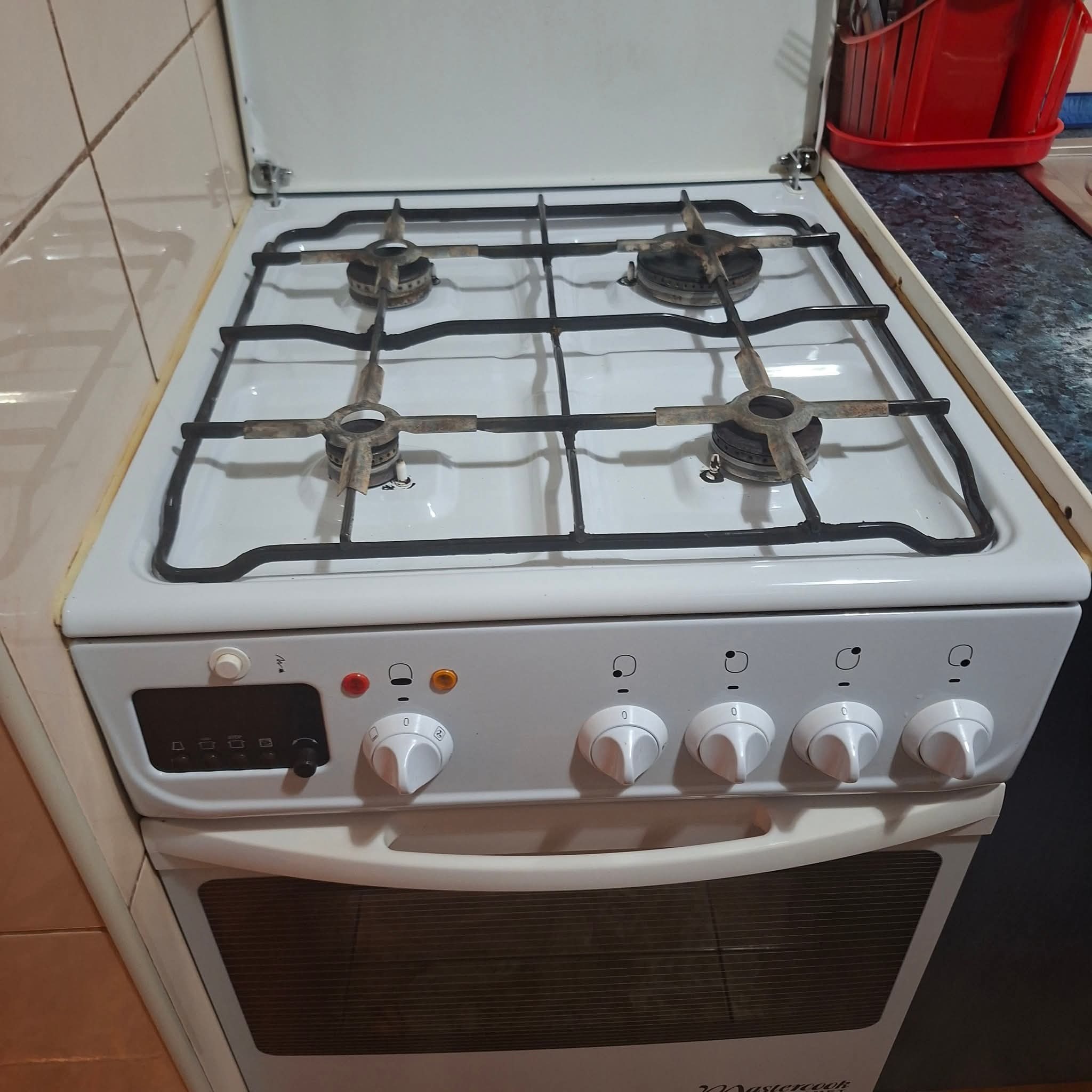 Biała kuchenka gazowa Mastercook z otwartą klapą, widoczne palniki i pokrętła. Na blacie resztki zabrudzeń. Tło stanowią kafelki i elementy wyposażenia kuchni.