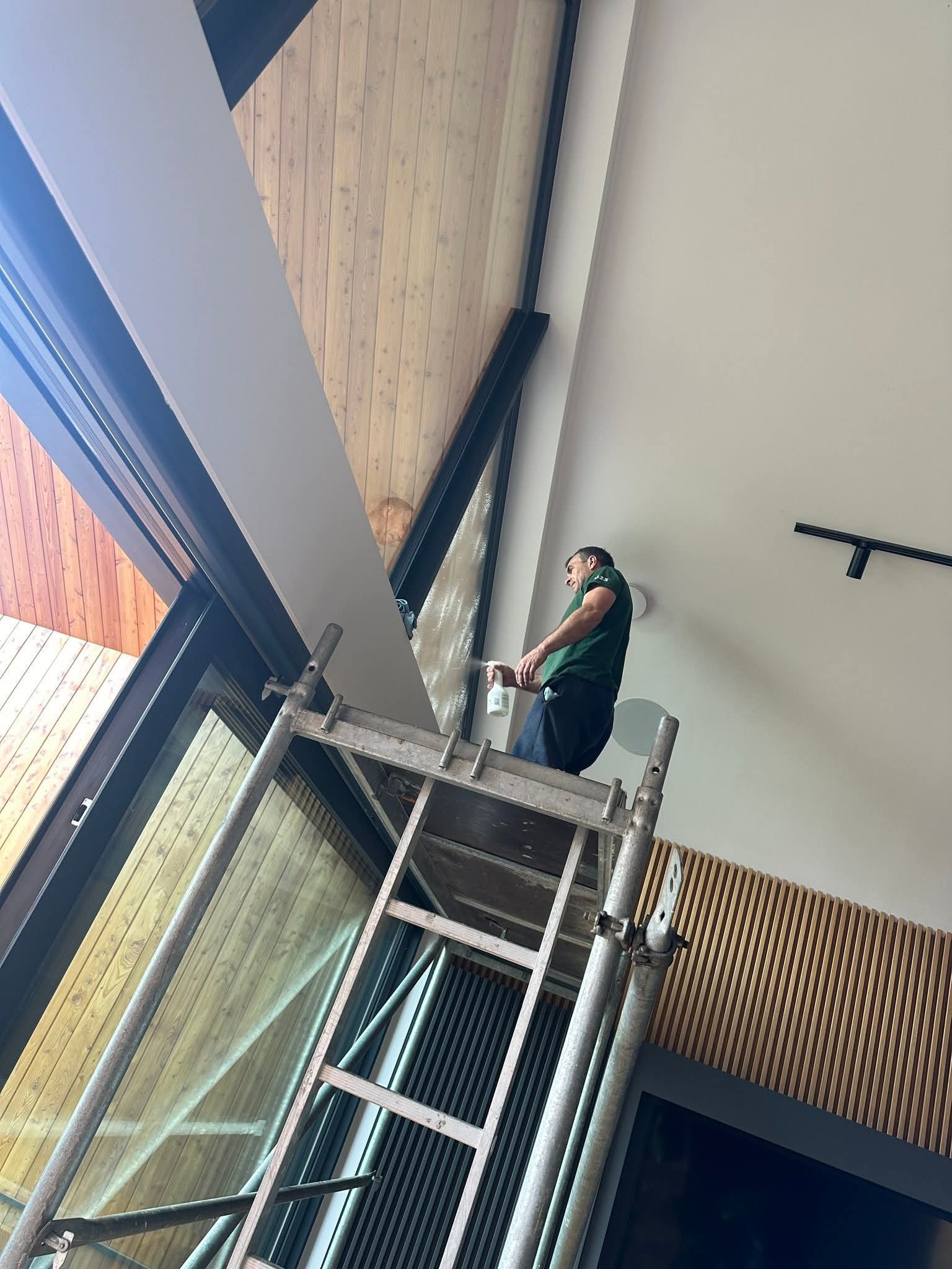 Mężczyzna na rusztowaniu czyści okno w nowoczesnym wnętrzu z drewnianą boazerią. Używa spryskiwacza, widok z dołu, okno o nietypowym kształcie.