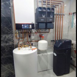 Marcin Hubert - Nowoczesna kotłownia z białym zasobnikiem, piecem gazowym Viessmann i rozbudowaną instalacją miedzianą na tle marmurowej ściany i płytek podłogowych.